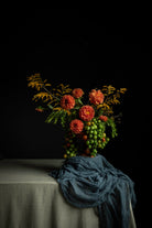 A still life floral arrangement with orange flowers, green leaves, and clusters of green and red berries, set on a draped table against a black background.