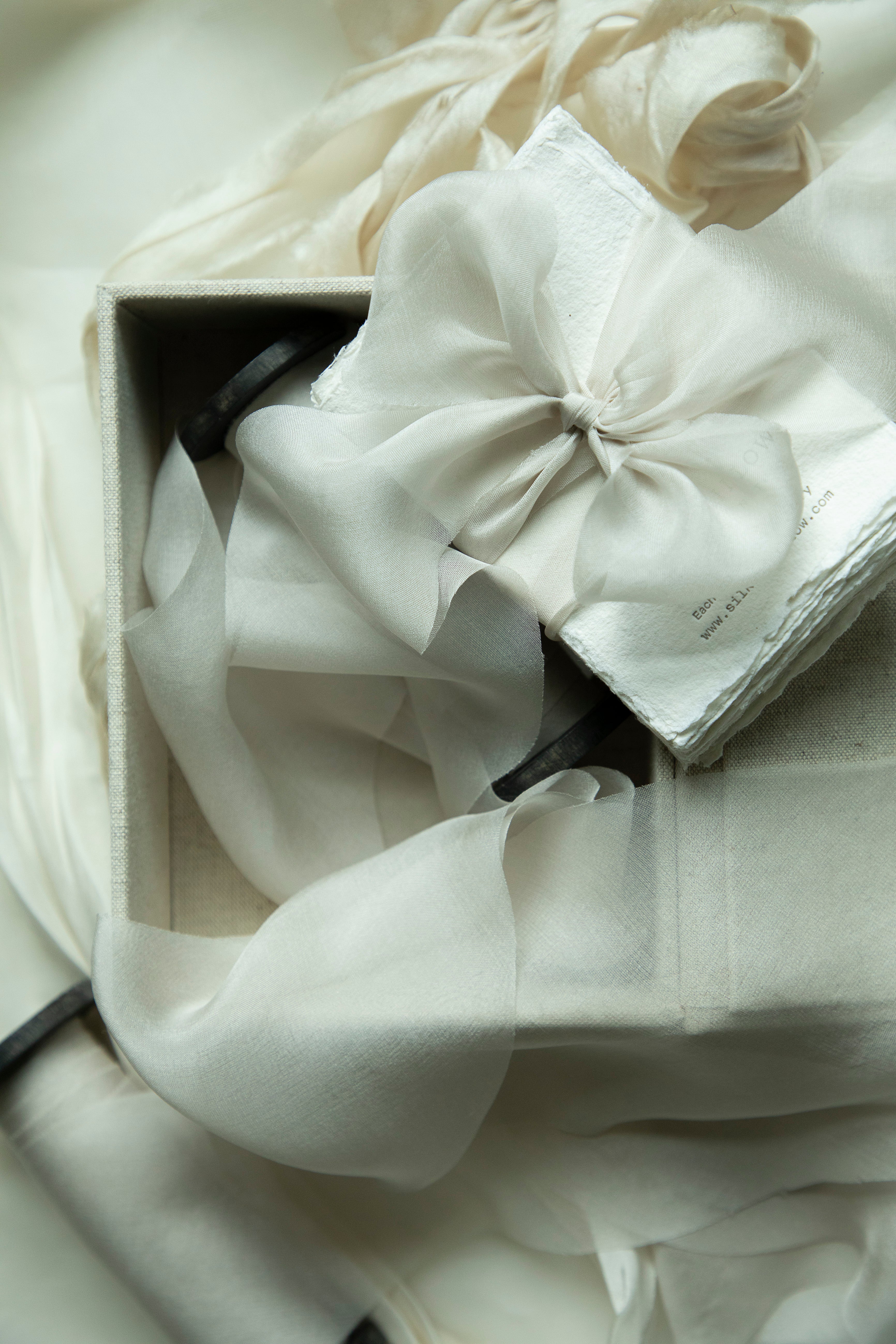 White fabric with a large bow in a box on a light background. A small stack of textured paper wrapped in a soft, pale ribbon, resting on a light fabric backdrop. 