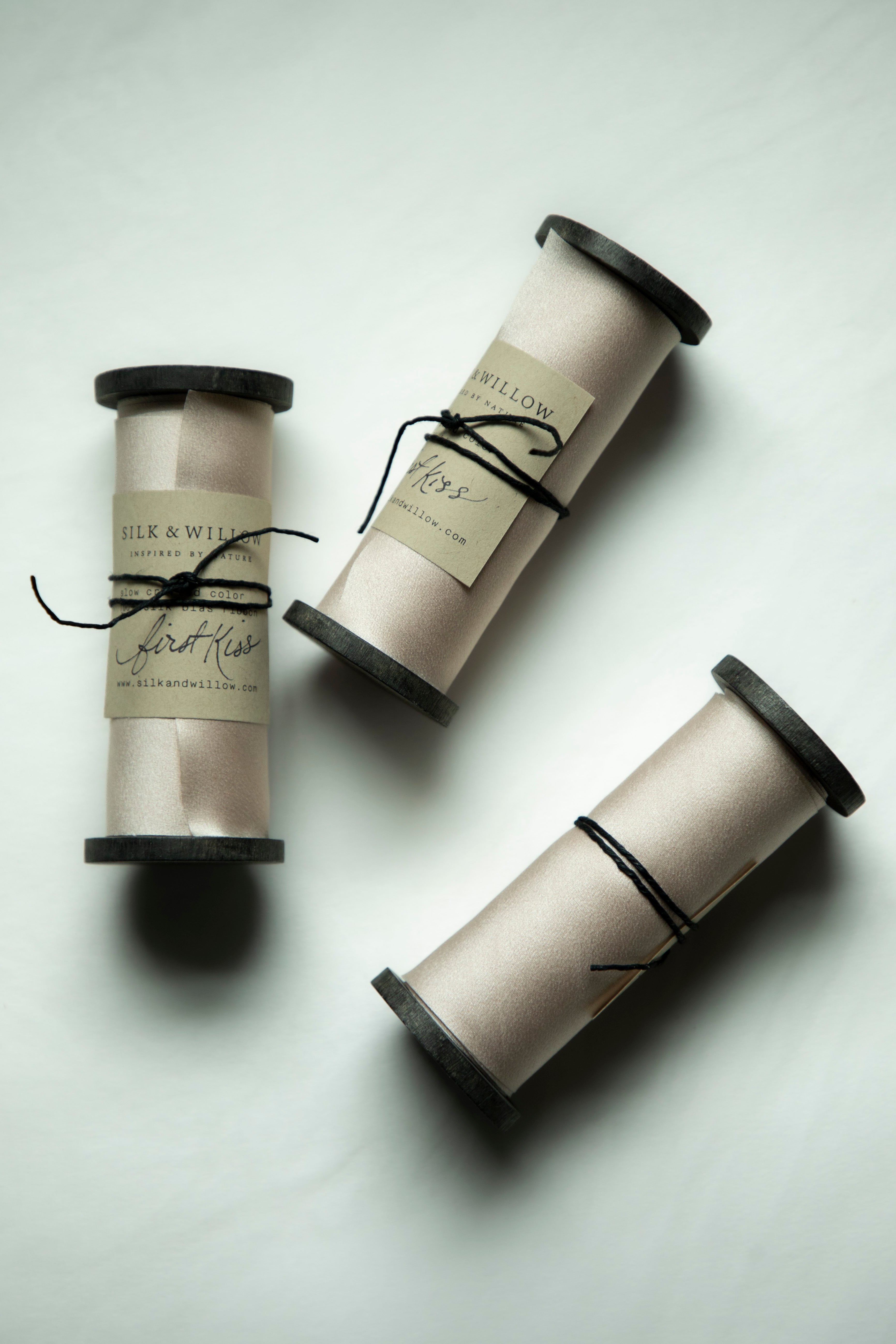 Three spools of silk ribbon in a light blush color on a black wood spool, tied with black string and featuring silk & Willow labels, arranged on a light gray background.