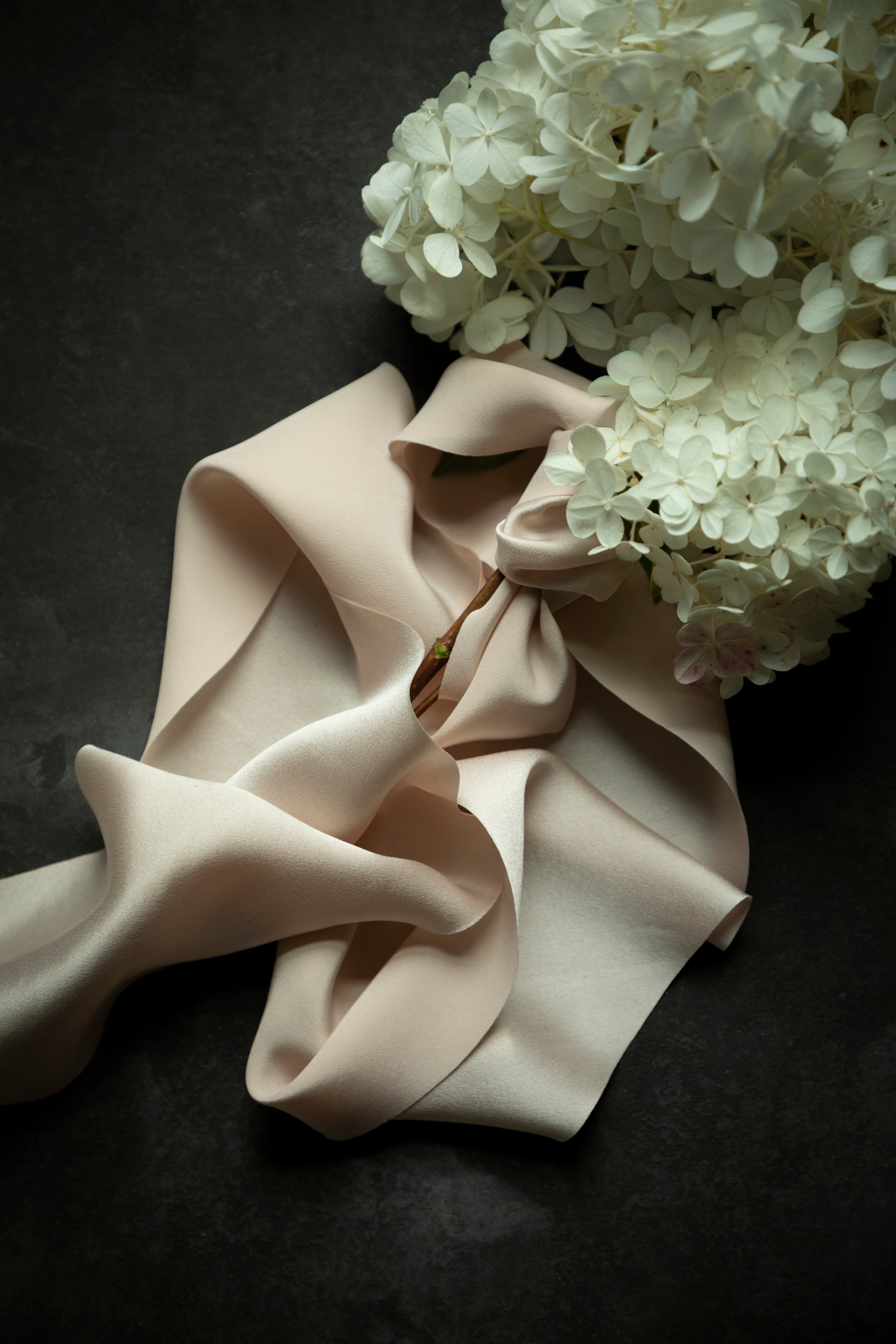 A draped piece of pale blush pink satin ribbon with soft folds, styled with white hydrangea flowers on a dark textured background.