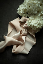 A draped piece of pale blush pink satin ribbon with soft folds, styled with white hydrangea flowers on a dark textured background.