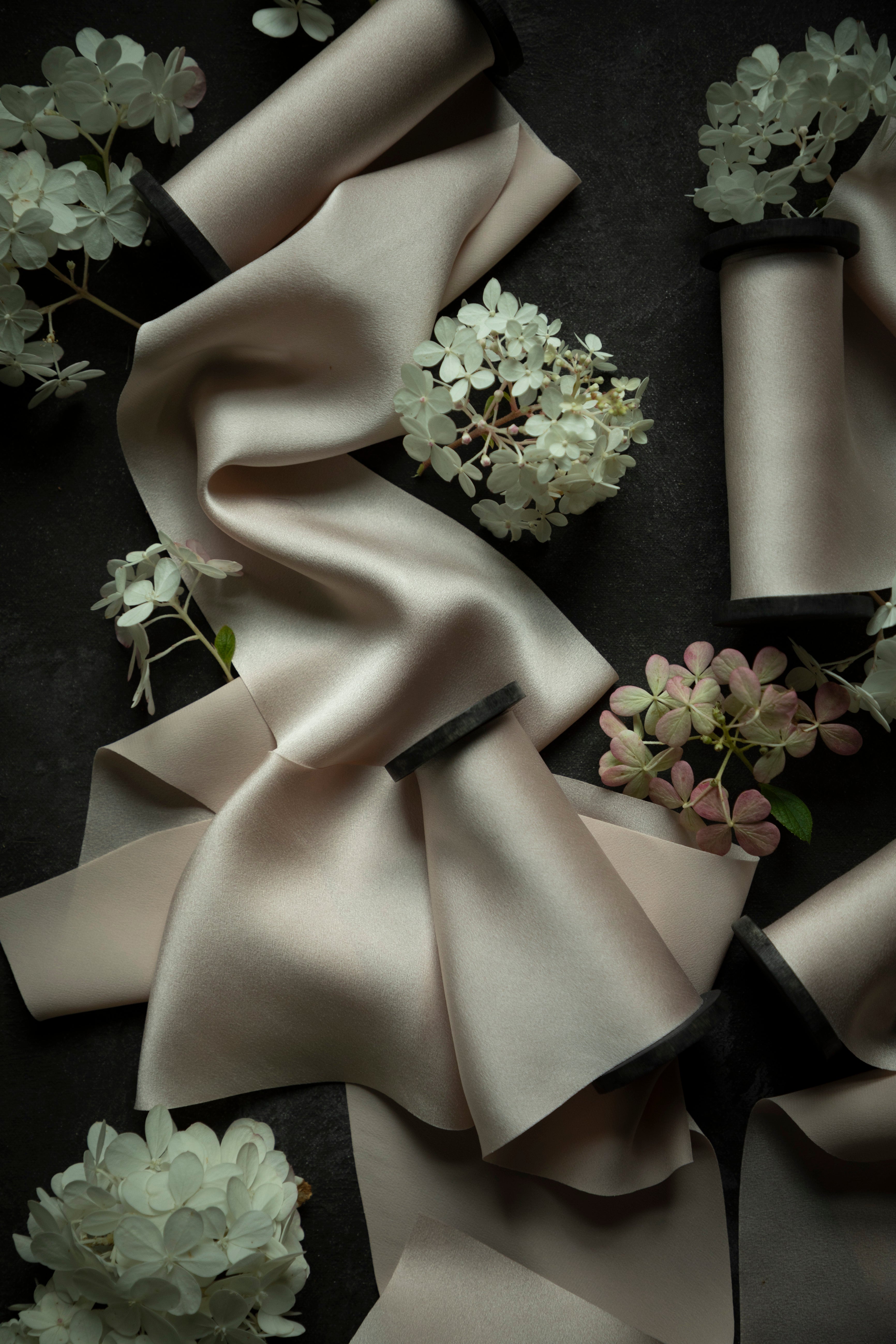 A draped piece of silk satin bridal bouquet ribbon in a light blush color, styled with white and pink hydrangea flowers on a dark textured background.