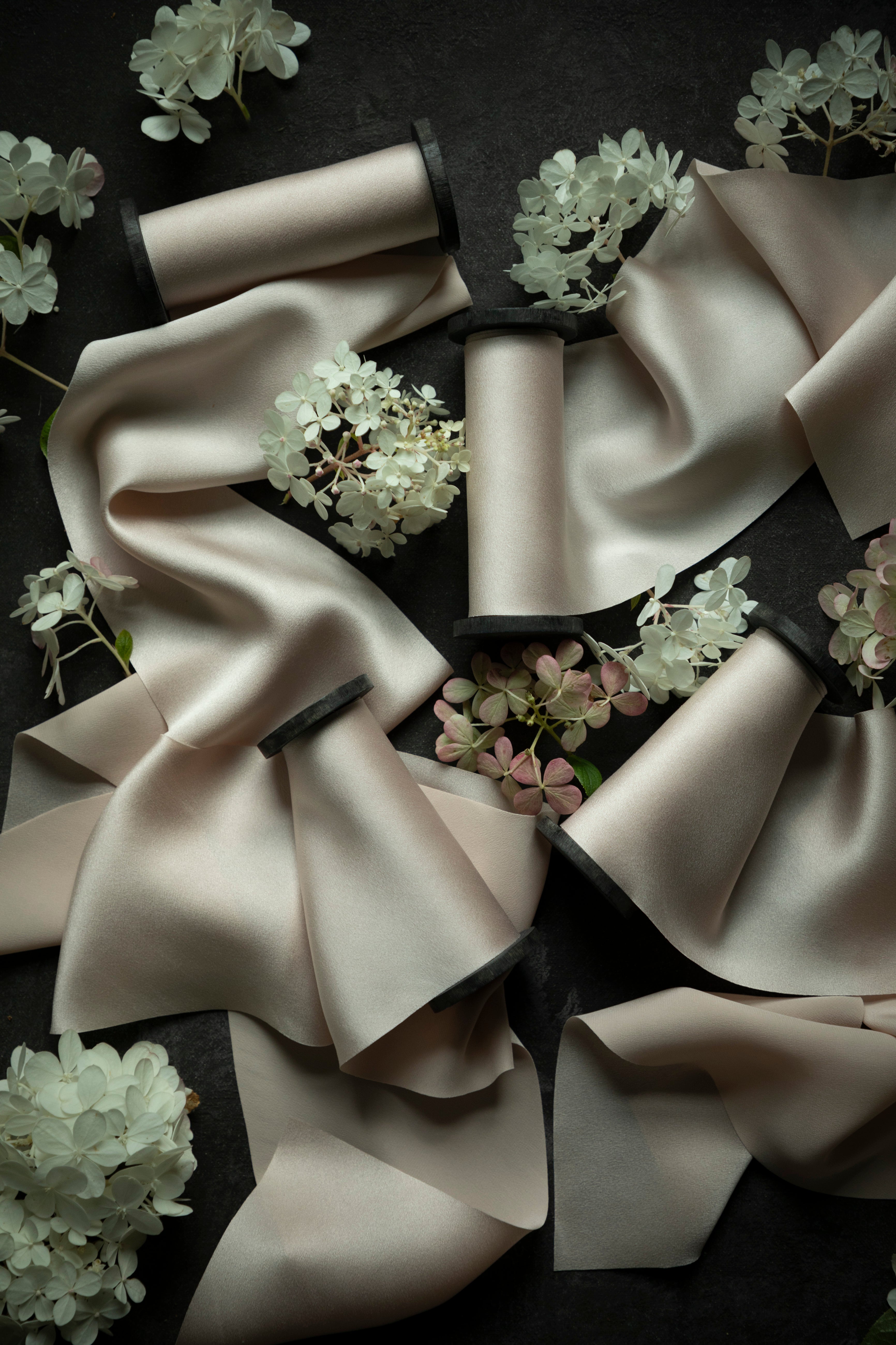 A styled flat-lay of multiple satin ribbon spools arranged on a dark background with decorative white and pale pink flowers. The ribbons are a warm blush-beige tone with dark inner ends visible on the spools.
