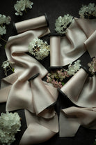 A styled flat-lay of multiple satin ribbon spools arranged on a dark background with decorative white and pale pink flowers. The ribbons are a warm blush-beige tone with dark inner ends visible on the spools.