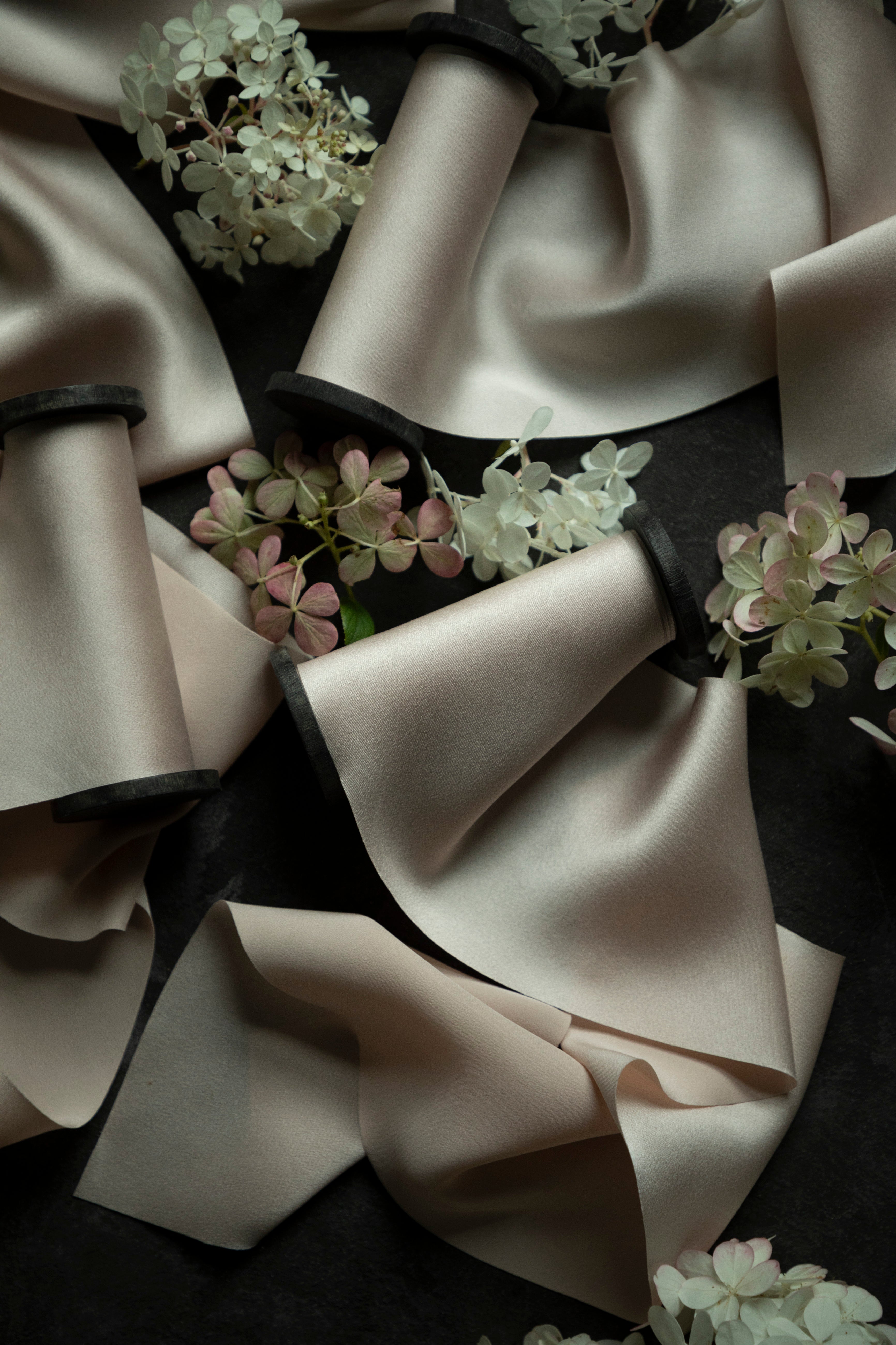Blush silk satin ribbon spools with black bands, arranged on a dark background with decorative white and pink hydrangea flowers.