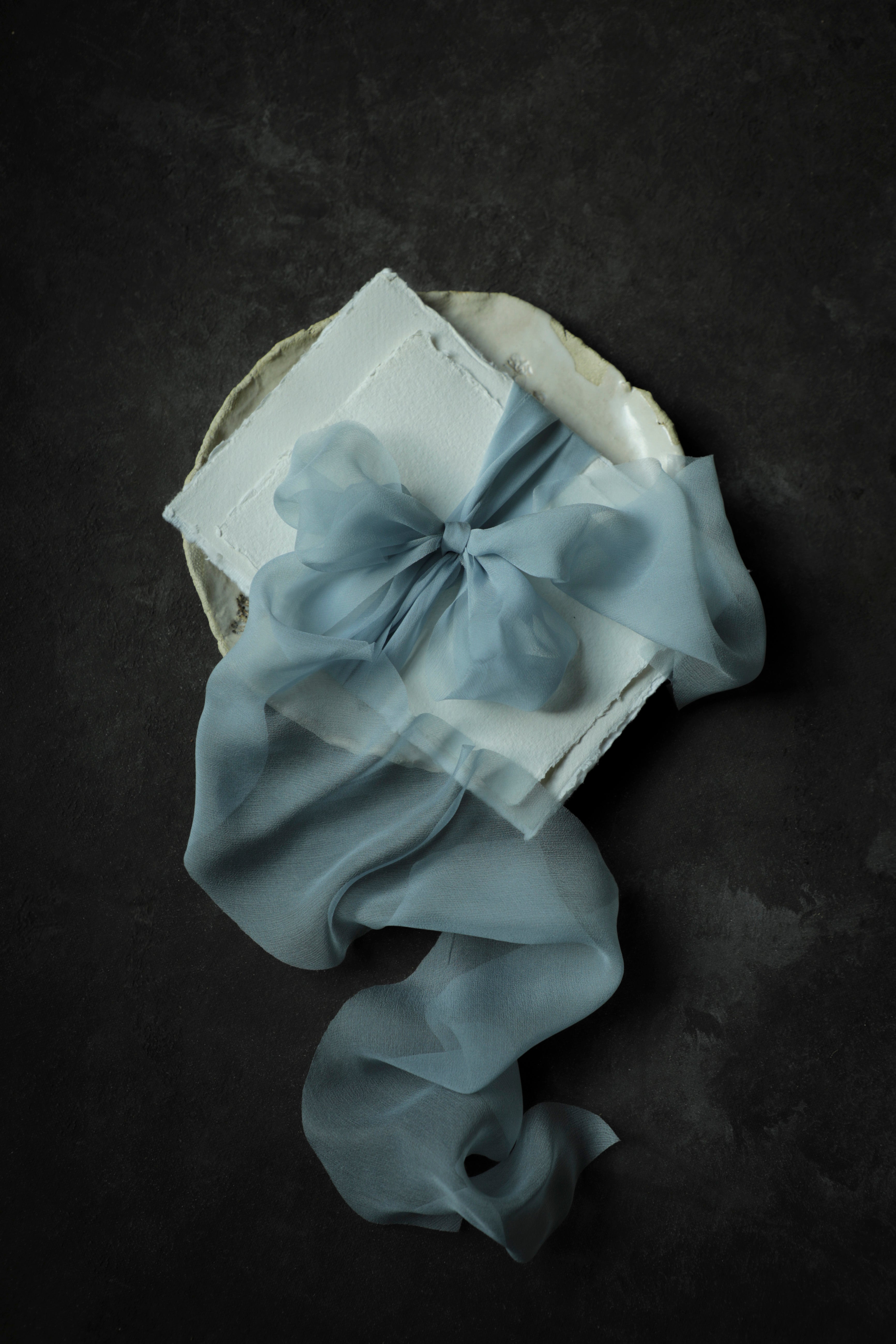 A light blue sheer organza ribbon tied into a large bow and draped over a white textured handmade deckle-edge paper envelope, presented on a dark textured background.
