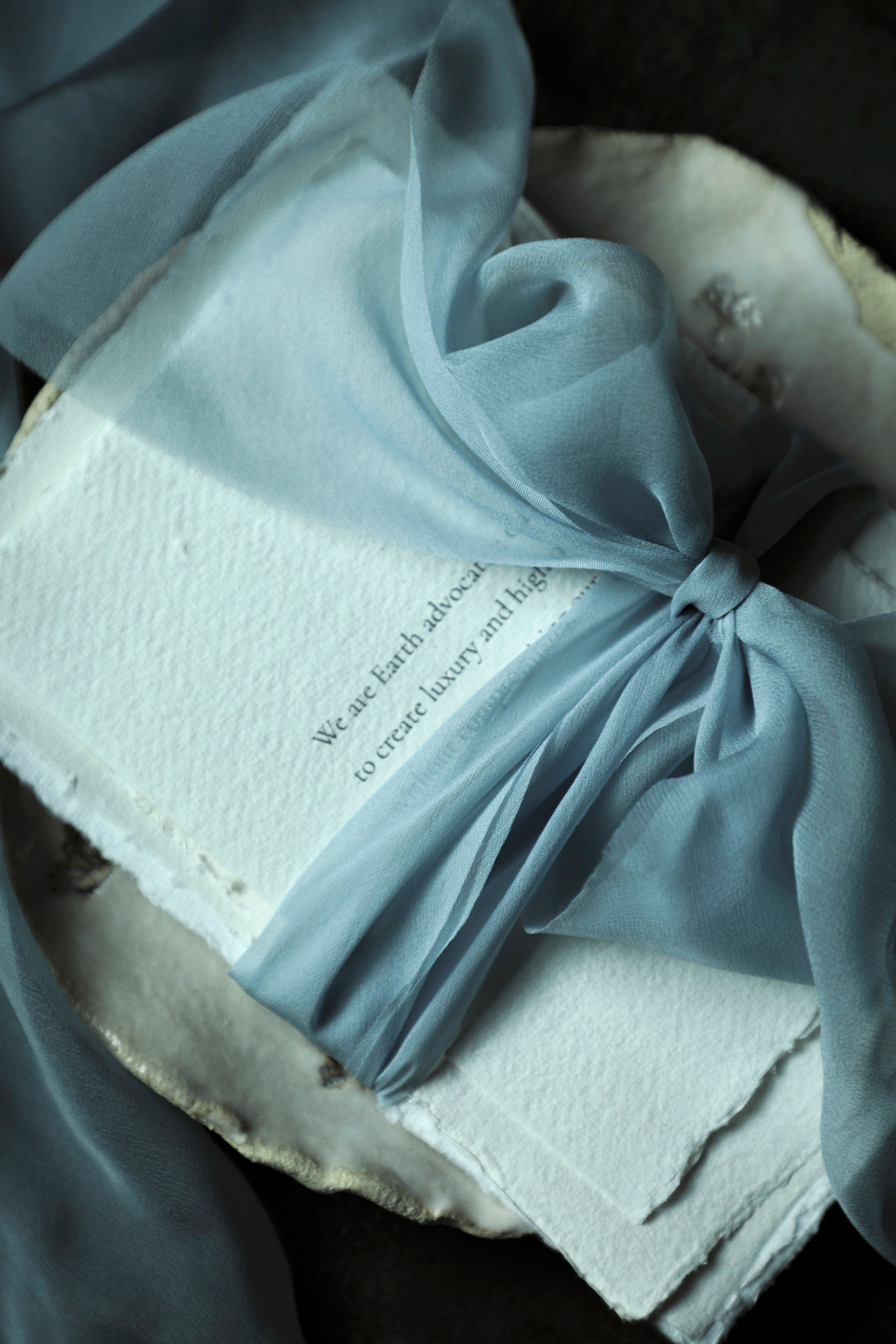 handmade deckled-edge paper wrapped with a frech blue sheer silk ribbon and tied with a big bow onto a handmade ceramic plate against a black backgraound.