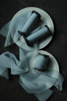 Three light blue spools of french blue silk ribbon arranged on white ceramic plates with sheer blue ribbons draped across a dark textured background.