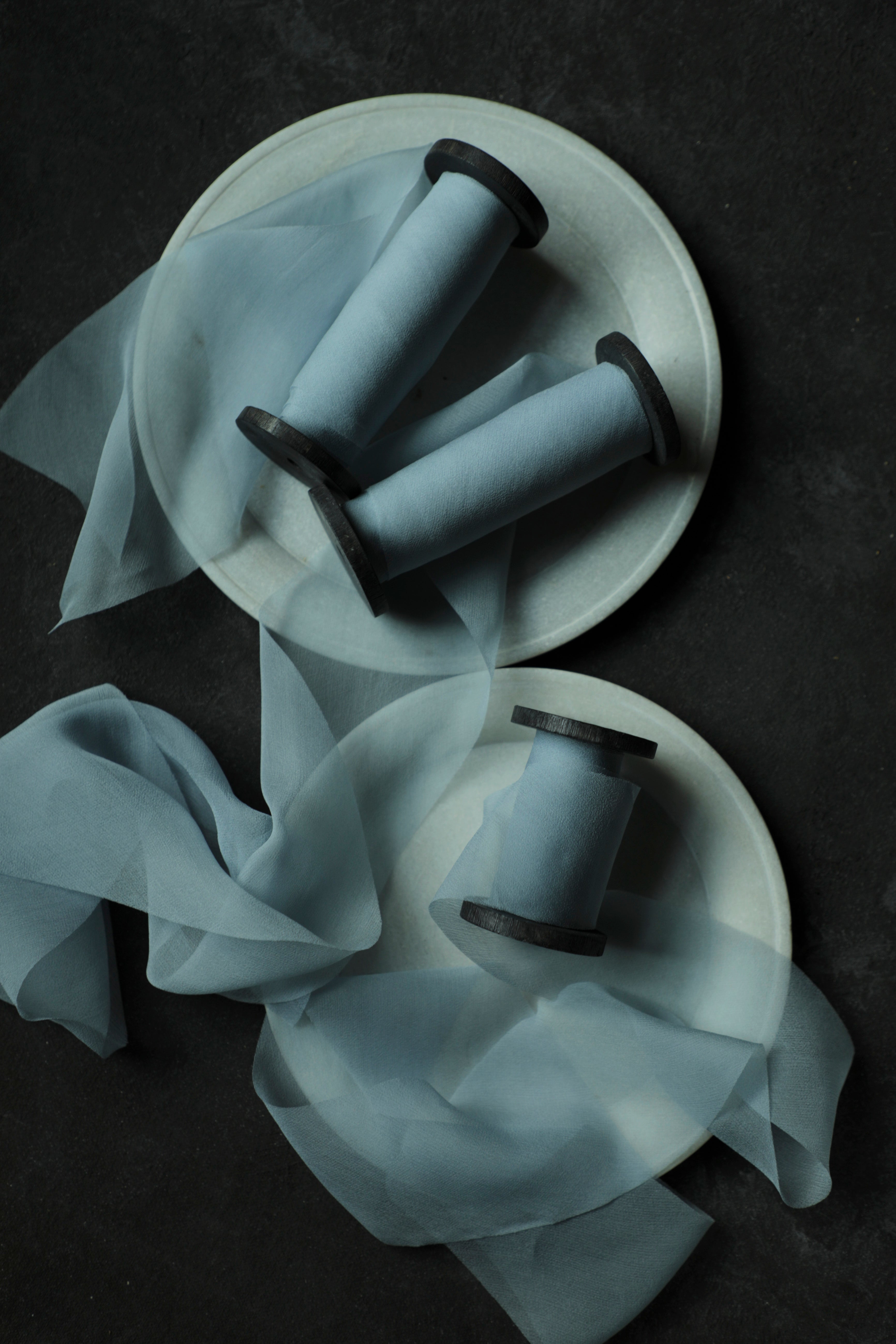 Three light blue spools of french blue silk ribbon arranged on white ceramic plates with sheer blue ribbons draped across a dark textured background.