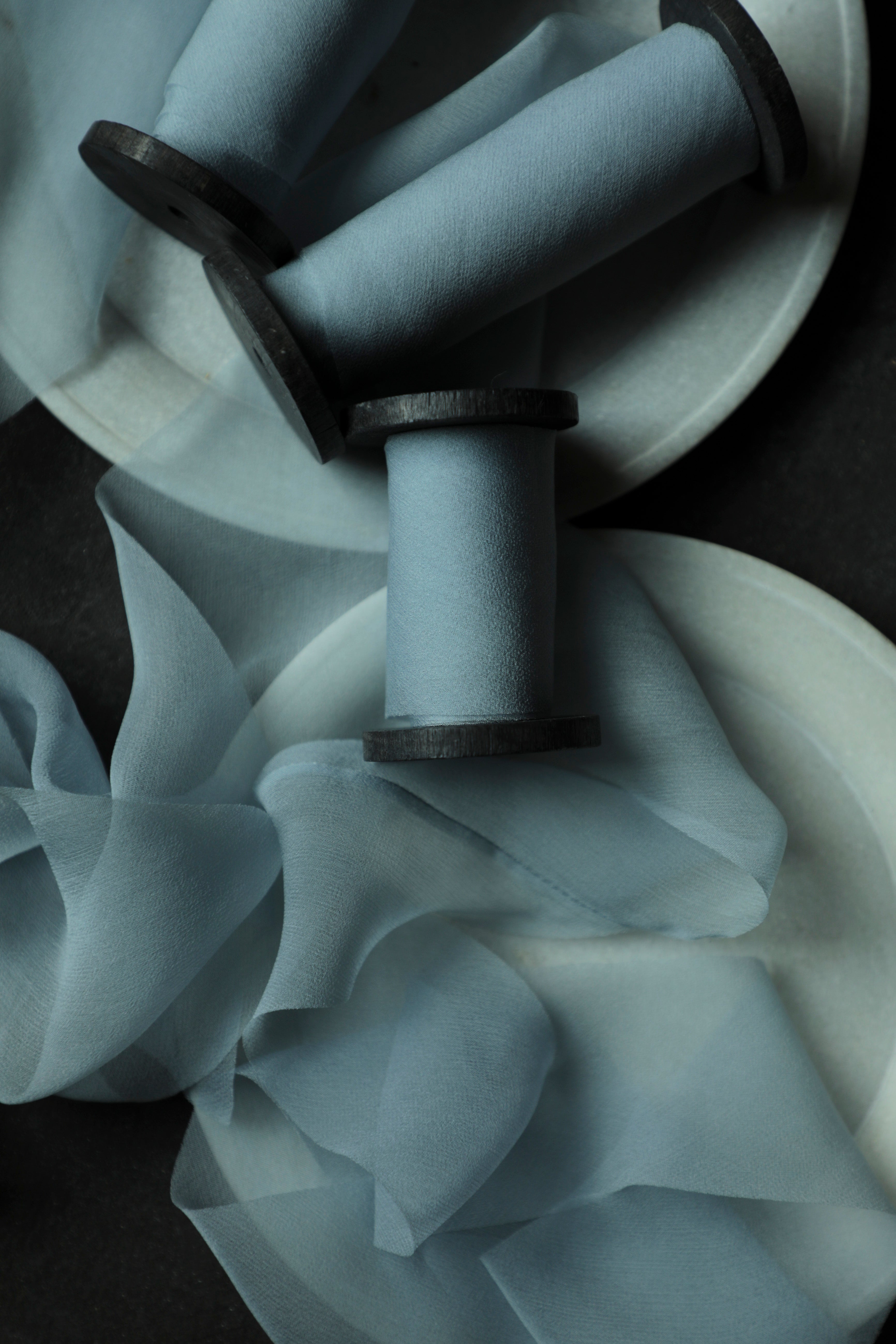 Light blue sheer silk ribbons draped over white circular plates, photographed in moody low light on a dark surface.