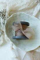 A small spool of sheer coral ribbon resting in a pale ceramic dish, surrounded by soft cream fabric and dried botanicals.