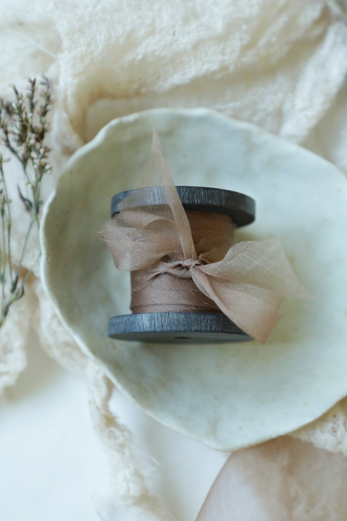 A small spool of sheer coral ribbon resting in a pale ceramic dish, surrounded by soft cream fabric and dried botanicals.