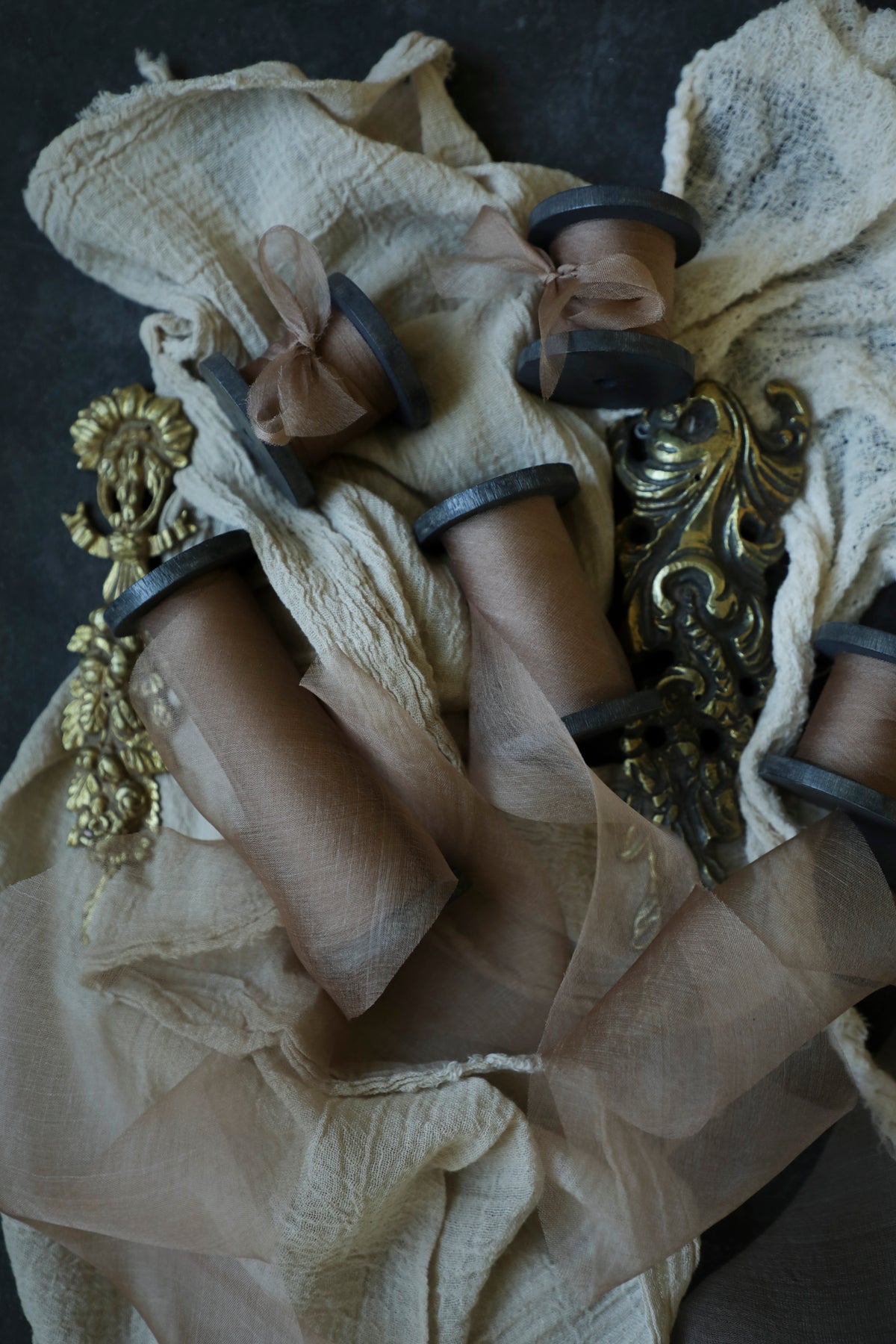 A styled flat-lay of vintage-style brown ribbon spools wrapped in sheer coral organza silk ribbon, accented with ornate gold baroque hardware on a dark textured background.