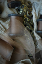 A close-up of sheer coral organza ribbon draped over a metallic gold ornate object, with cream lace fabric in the background.
