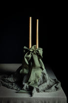 A decorative centerpiece featuring two tall natural wood taper candles standing upright in a gathered olive green fabric base with a large bow, draped over a textured gray fabric on a dark table against a black background.