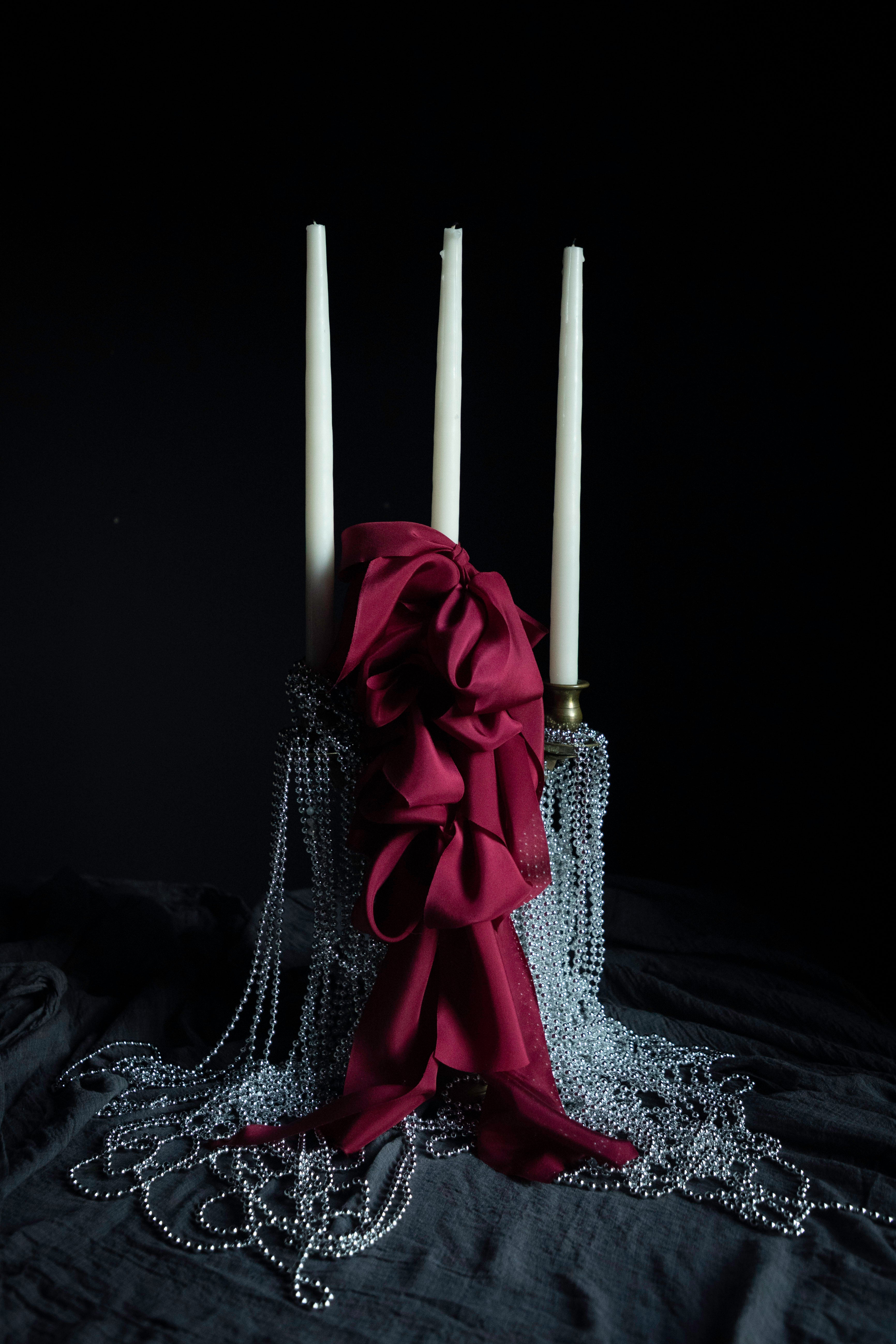 A gothic-style three taper candle holder with white candles, a large dark burgundy satin bow, and cascading silver bead chains on a black fabric background.