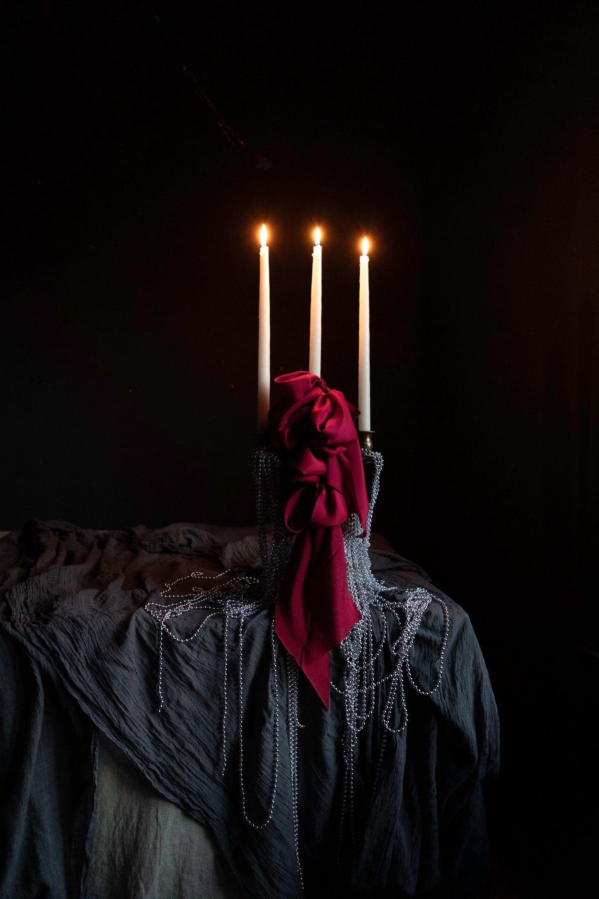 candles draped with metal beads and red ribbon bows on layered gray table textiles against a dark background