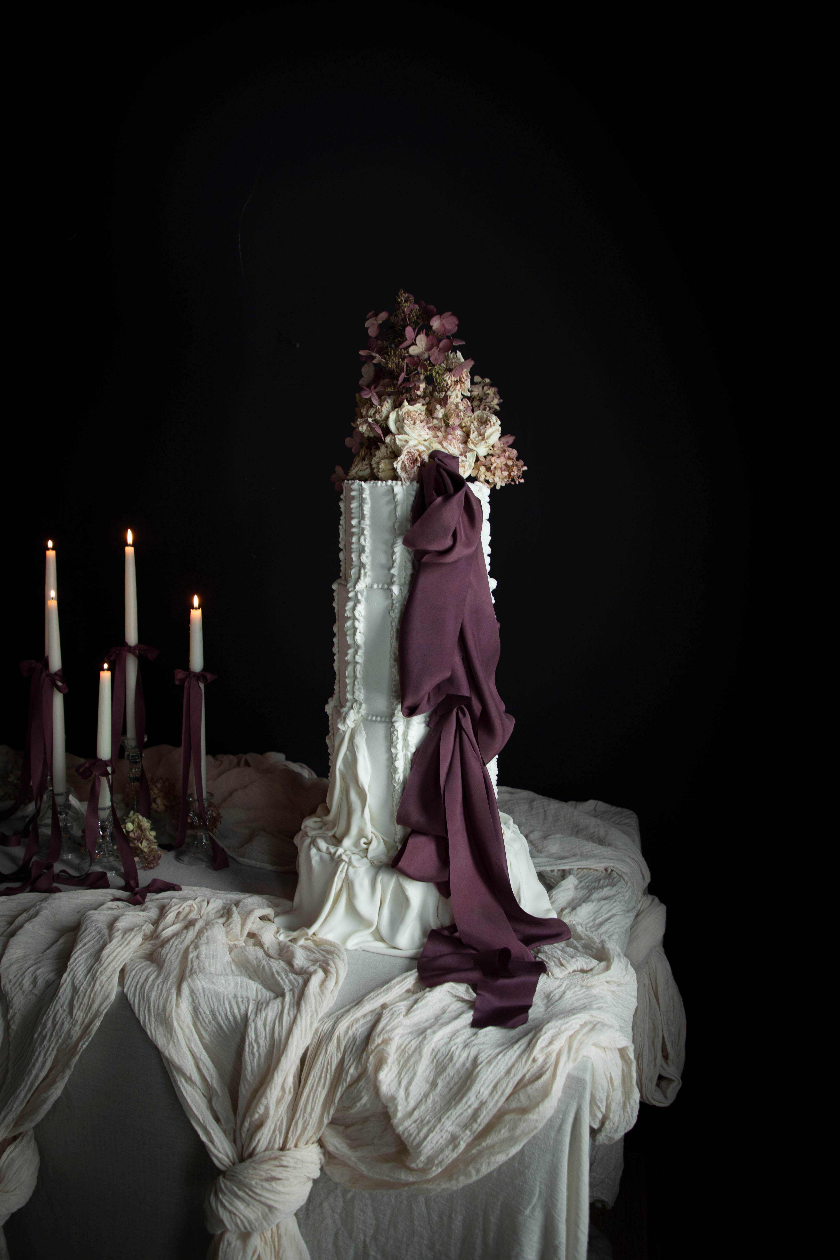 White wedding cake with purple noir ribbon decor on a table against a dark background