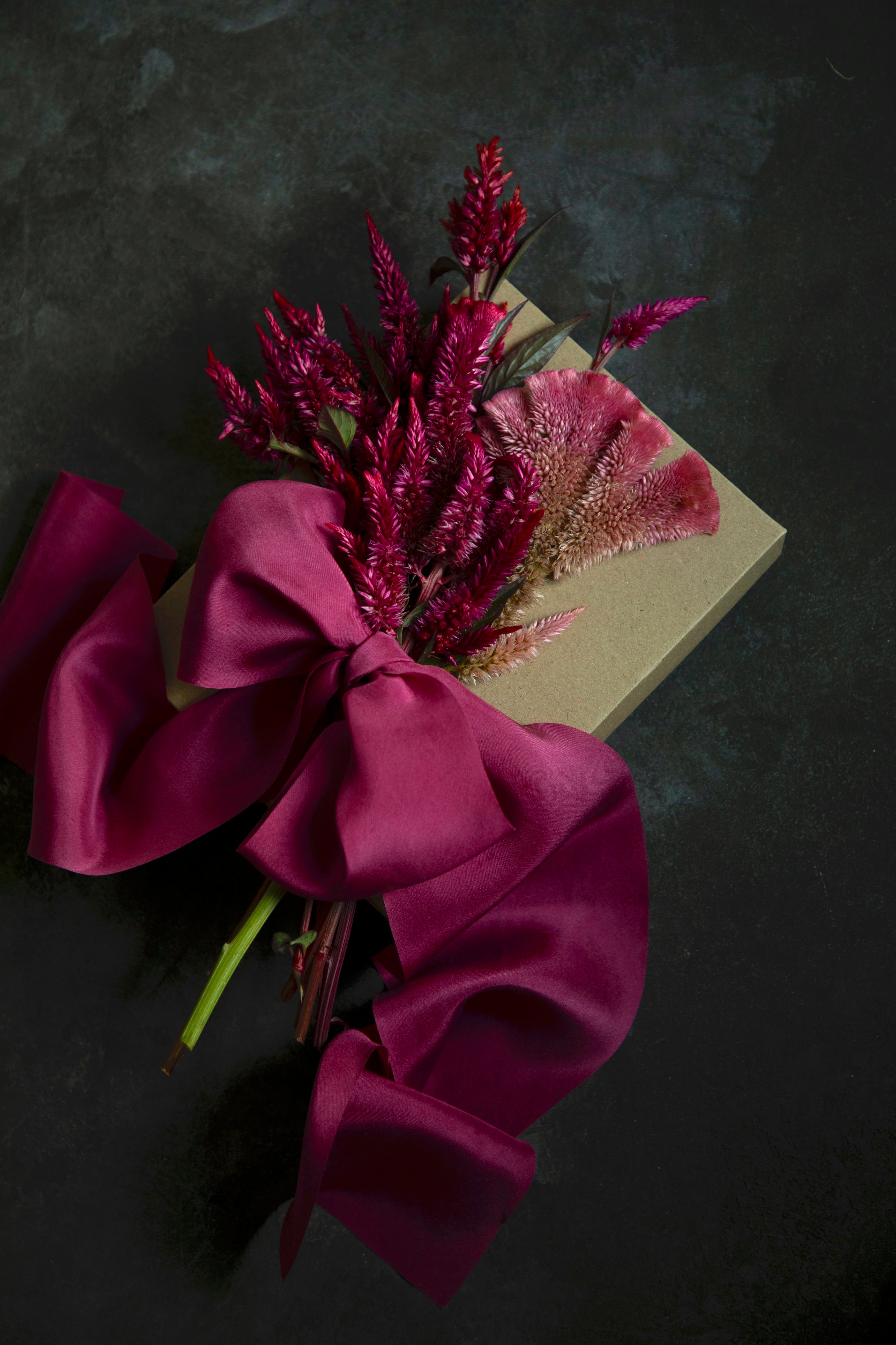 A dried floral arrangement featuring deep magenta dried flowers, a pinkish-purple feather grass flower, and a large magenta satin bow wrapped around a square kraft paper box, set against a dark textured background.