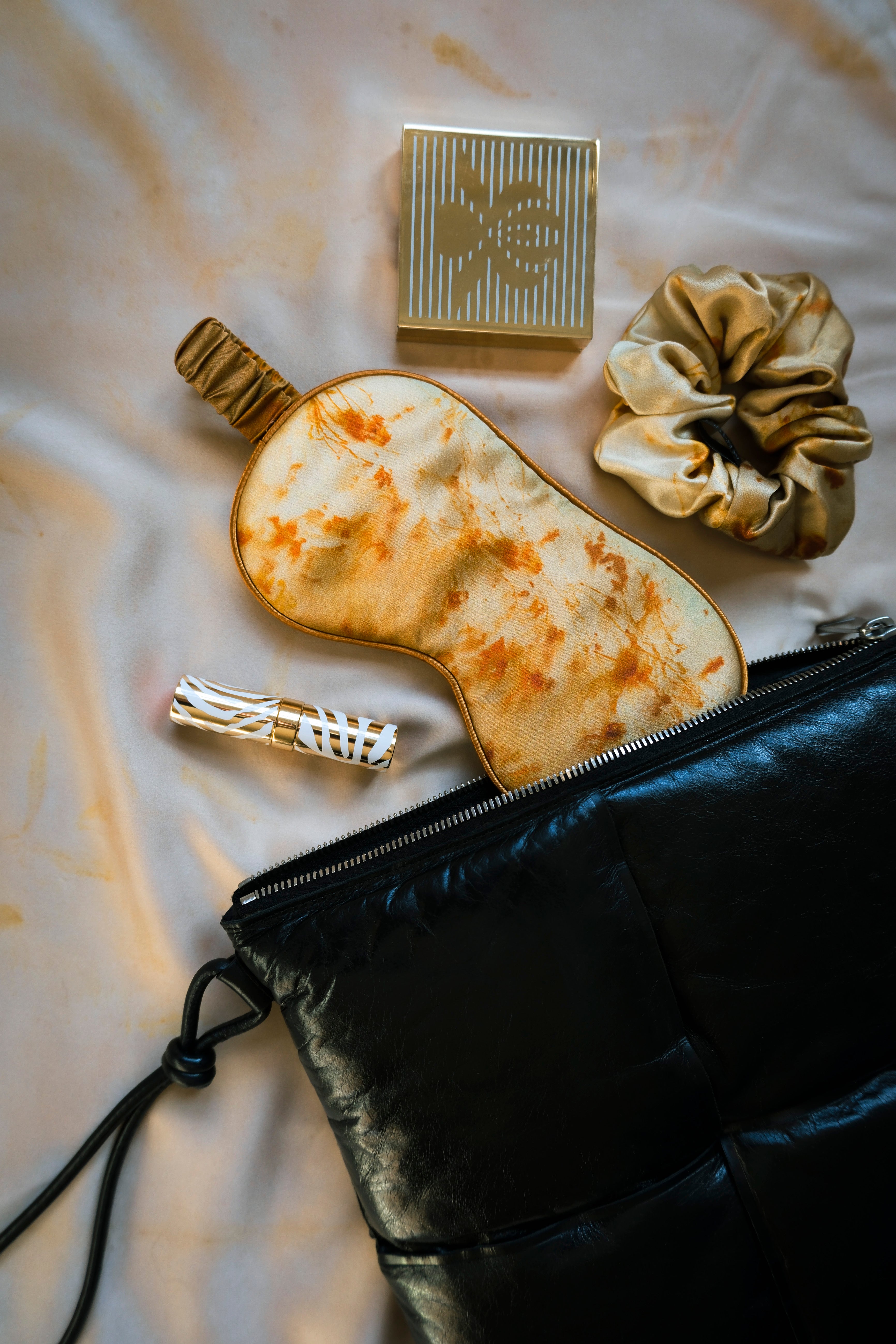 travel essentials, a silk Sleep mask, a sisley lip gloss, and a hair scrunchie on a silk surface with a black bag.