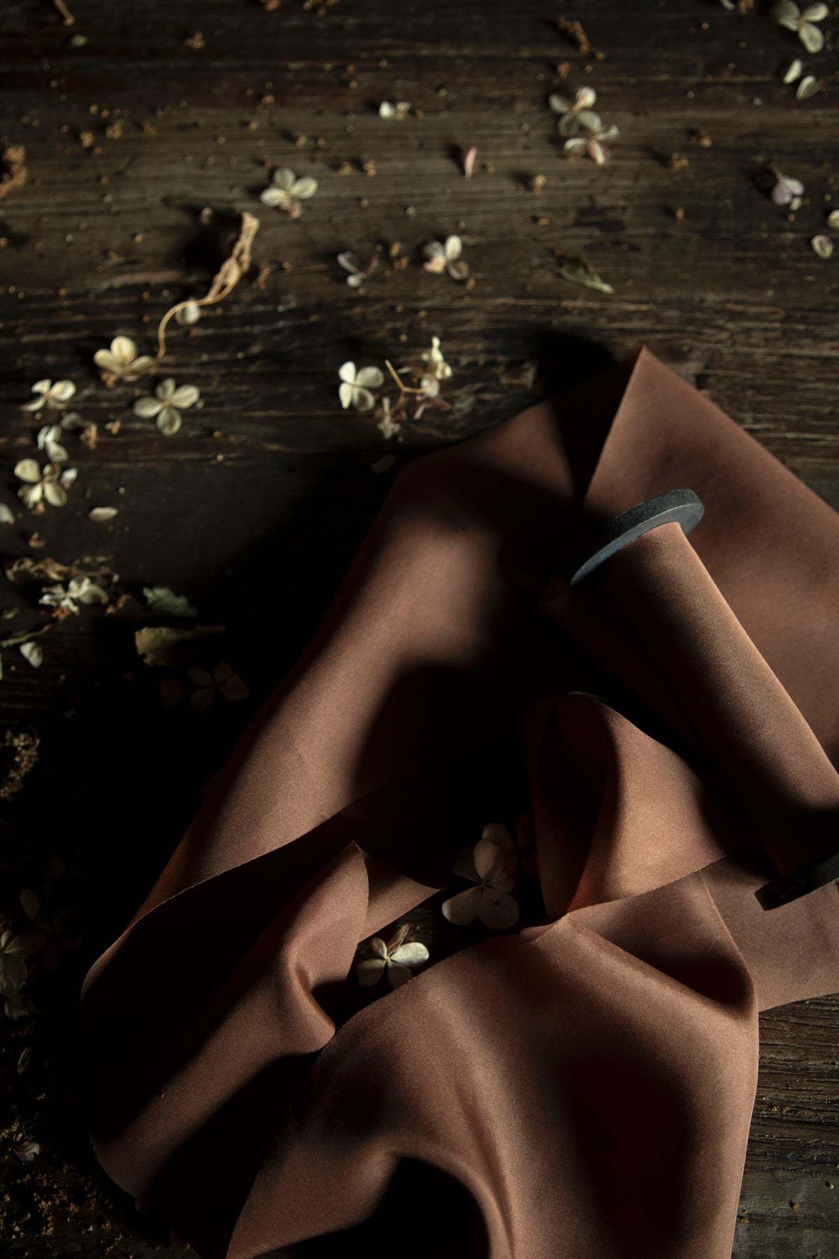 A close-up of draped brown ribbon on a wooden spool, arranged on a dark rustic wooden surface with scattered small white flowers.
