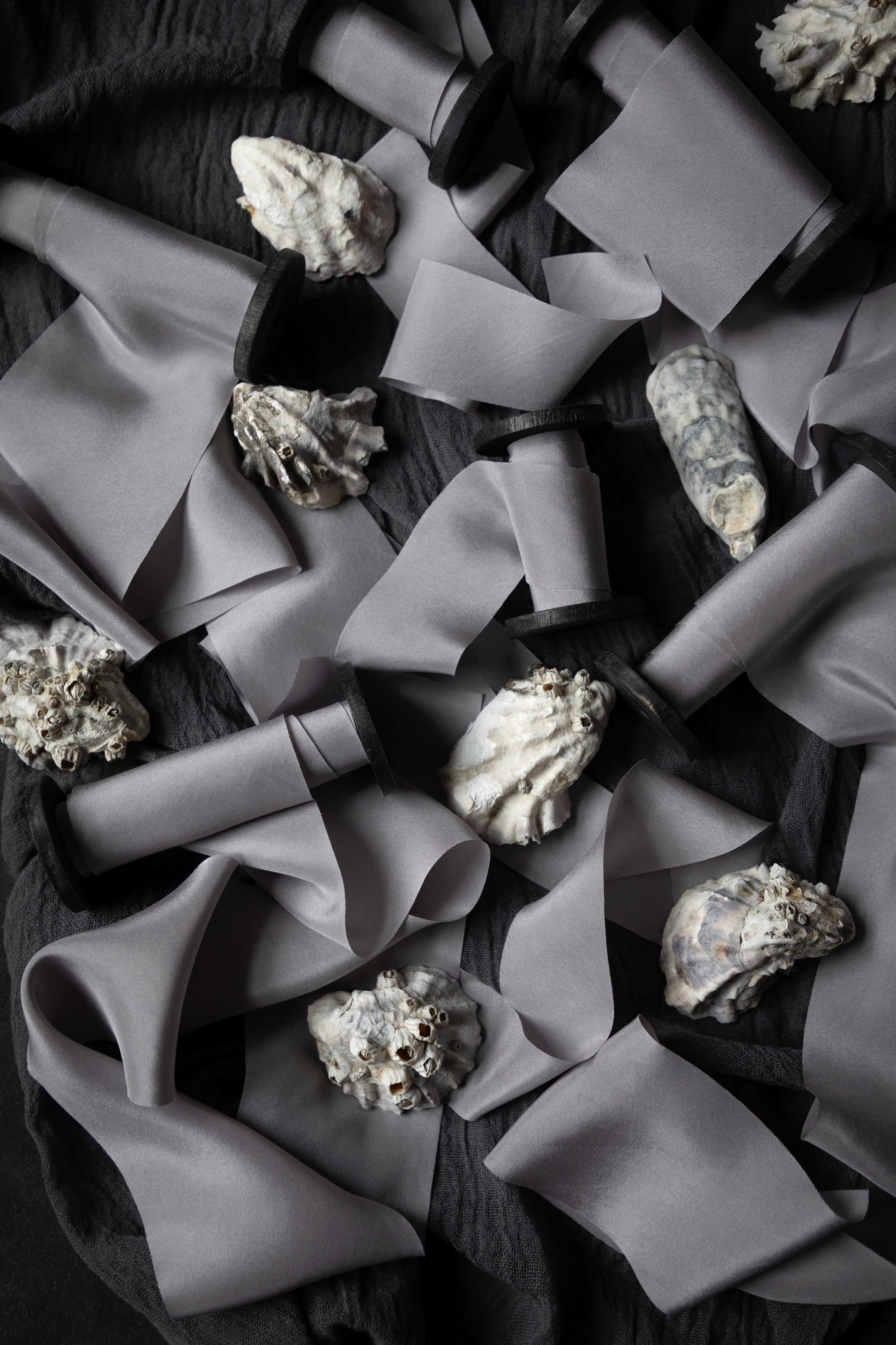 A styled flat-lay of gray silk ribbon scattered across a dark textured surface, interspersed with small white and gray seashells.