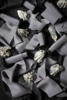 A styled flat-lay of gray silk ribbon scattered across a dark textured surface, interspersed with small white and gray seashells.