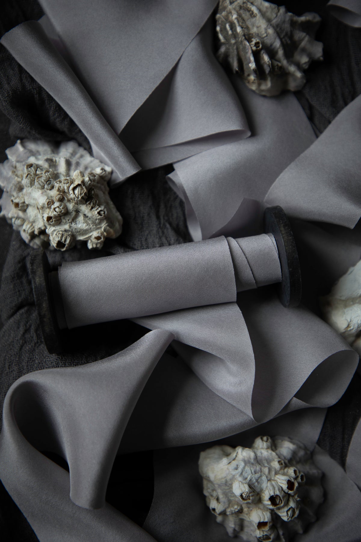 A close-up of draped gray silk ribbon arranged on a dark textured background, with small shell-like decorative props scattered around.