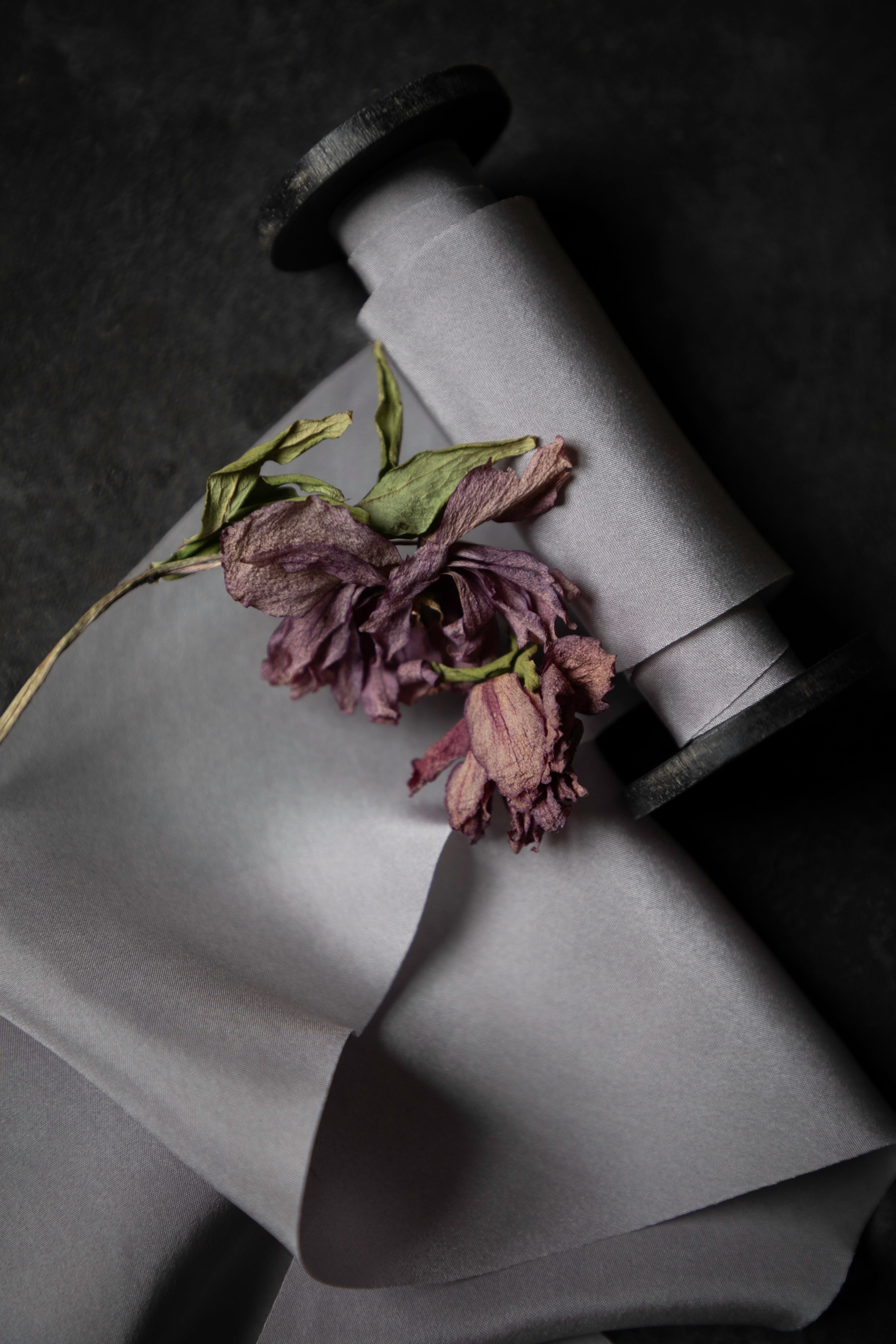 A roll of light gray silk ribbon with a dried purple flower resting on top, photographed on a dark textured background.