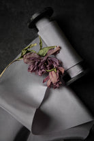 A roll of light gray silk ribbon with a dried purple flower resting on top, photographed on a dark textured background.