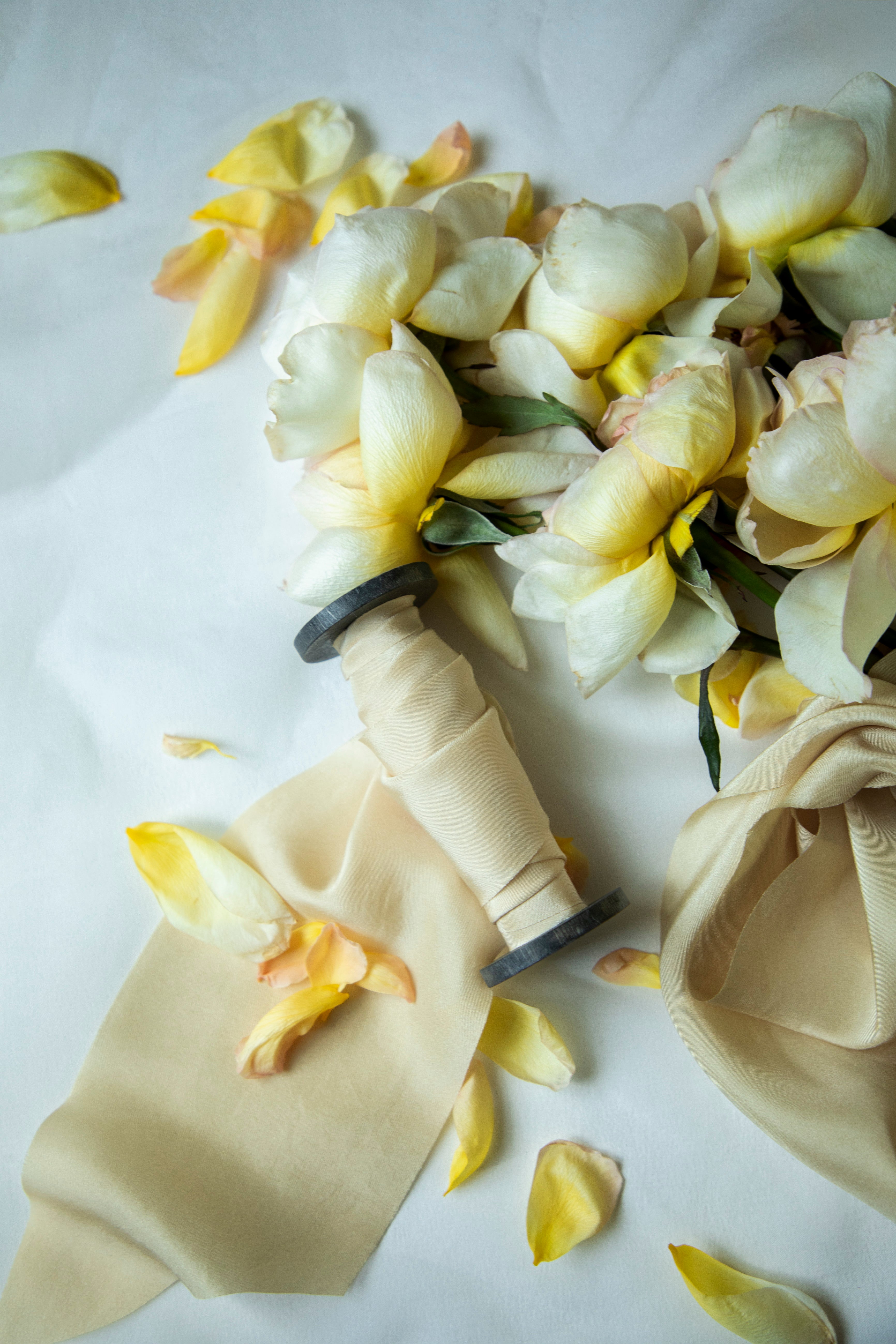 A bouquet of pale yellow and white flowers arranged on a white surface with a cream satin ribbon and scattered flower petals.