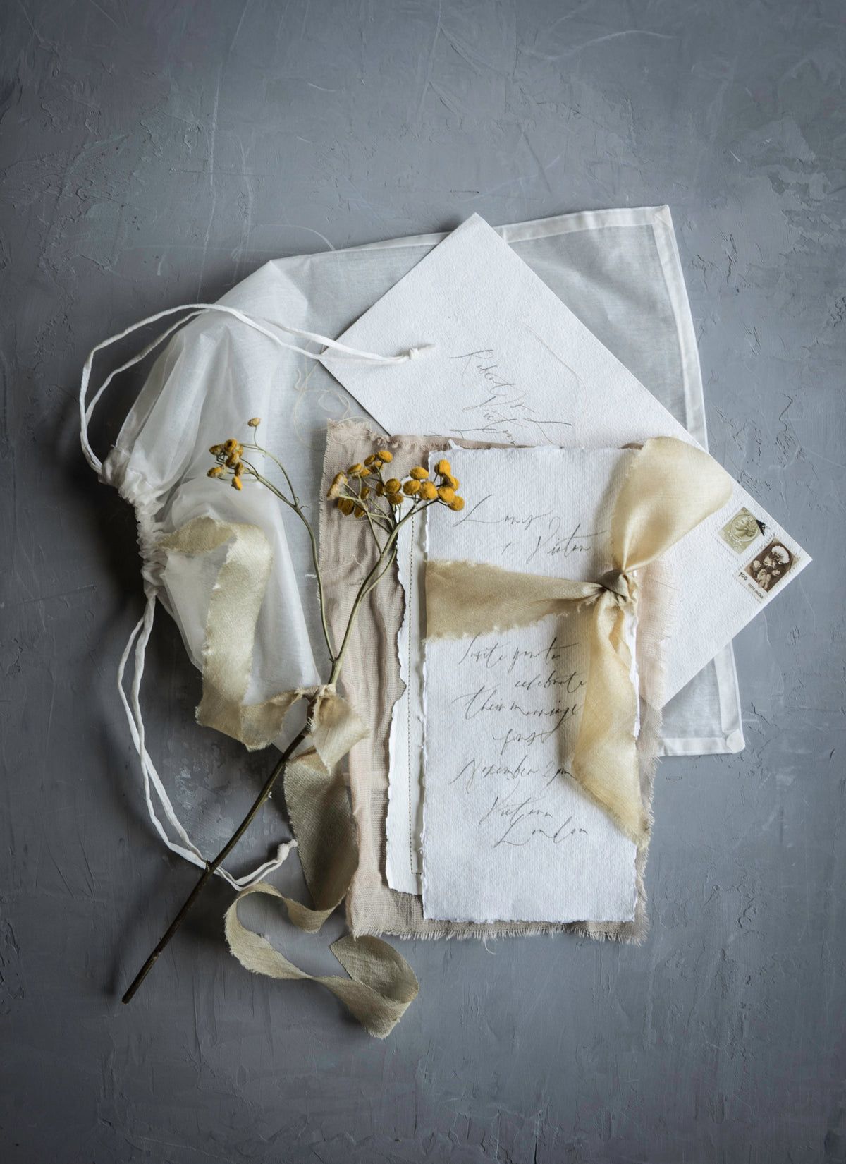 A styled flat-lay of vintage-inspired stationery on a textured gray background, featuring a sheer white drawstring pouch, a cream silk ribbon, a piece of raw-edge paper with cursive writing, a vintage-style postcard, and small yellow dried flowers.