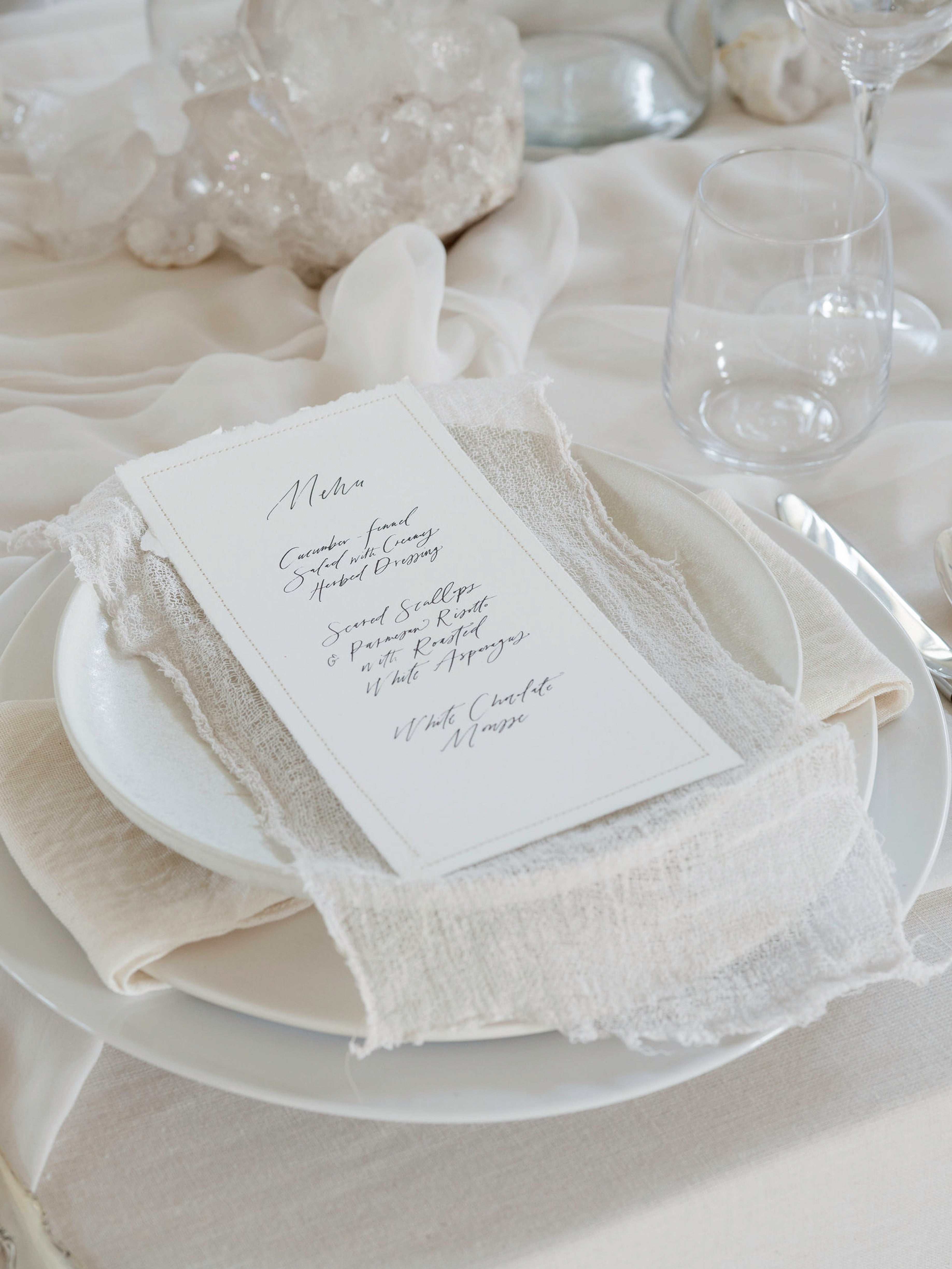 A wedding place card with elegant black script lettering, styled on a white plate with a cream textured linen napkin and matching tablecloth.