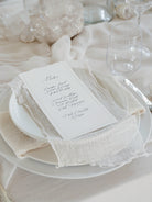 A wedding place card with elegant black script lettering, styled on a white plate with a cream textured linen napkin and matching tablecloth.
