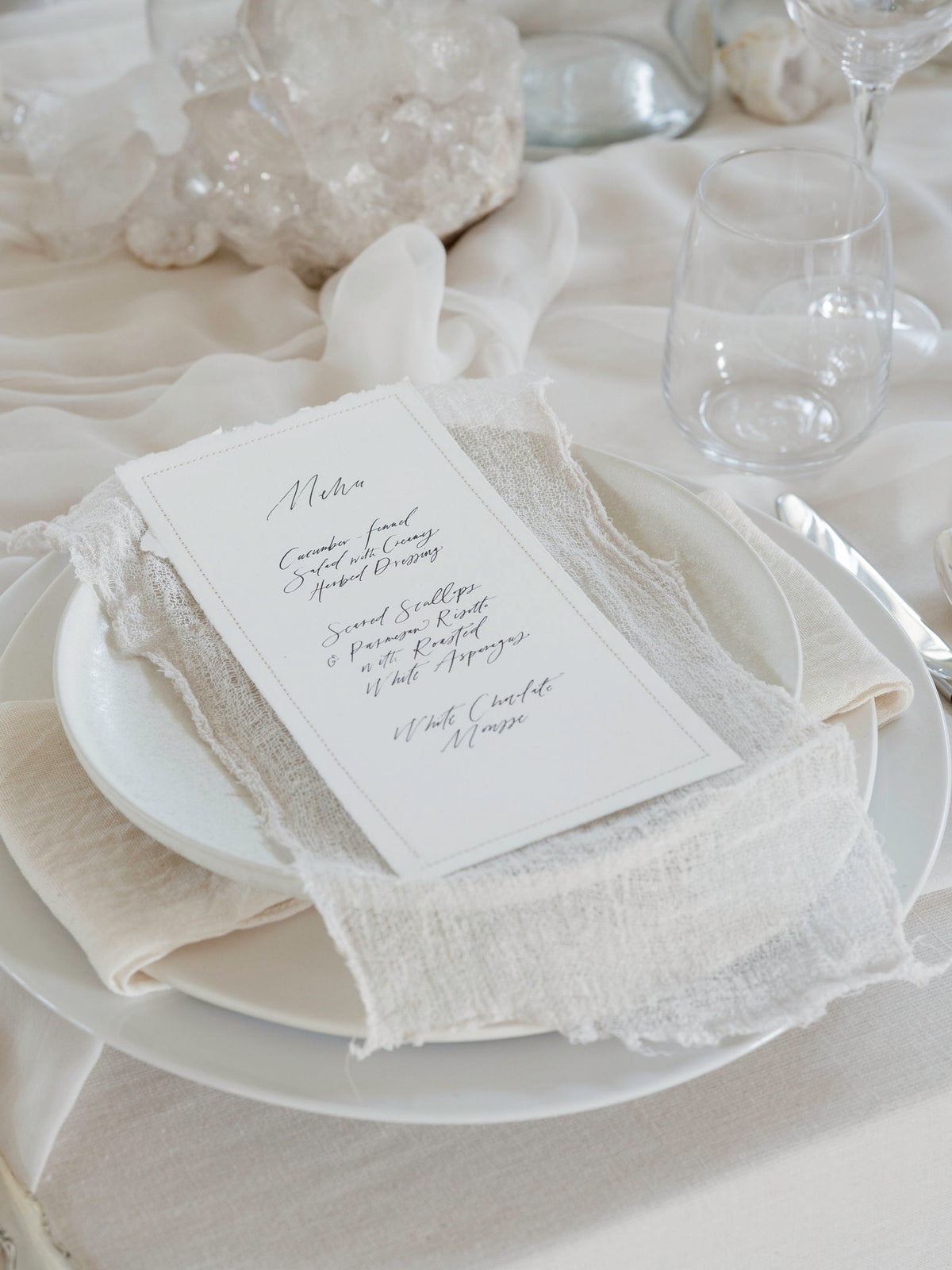 A wedding place card with elegant black script lettering, styled on a white plate with a cream textured linen napkin and matching tablecloth.