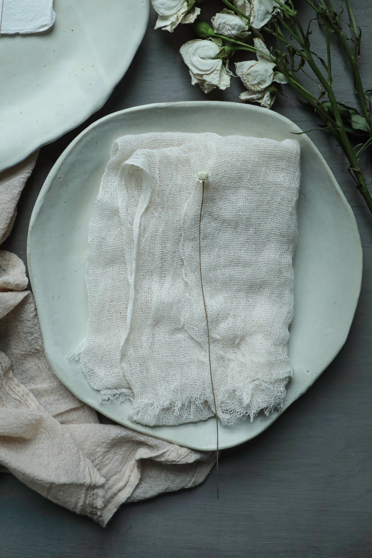 A folded off-white linen cloth napkin with frayed edges rests on a round ceramic plate, styled with dried white roses and neutral fabric on a dark gray tabletop.