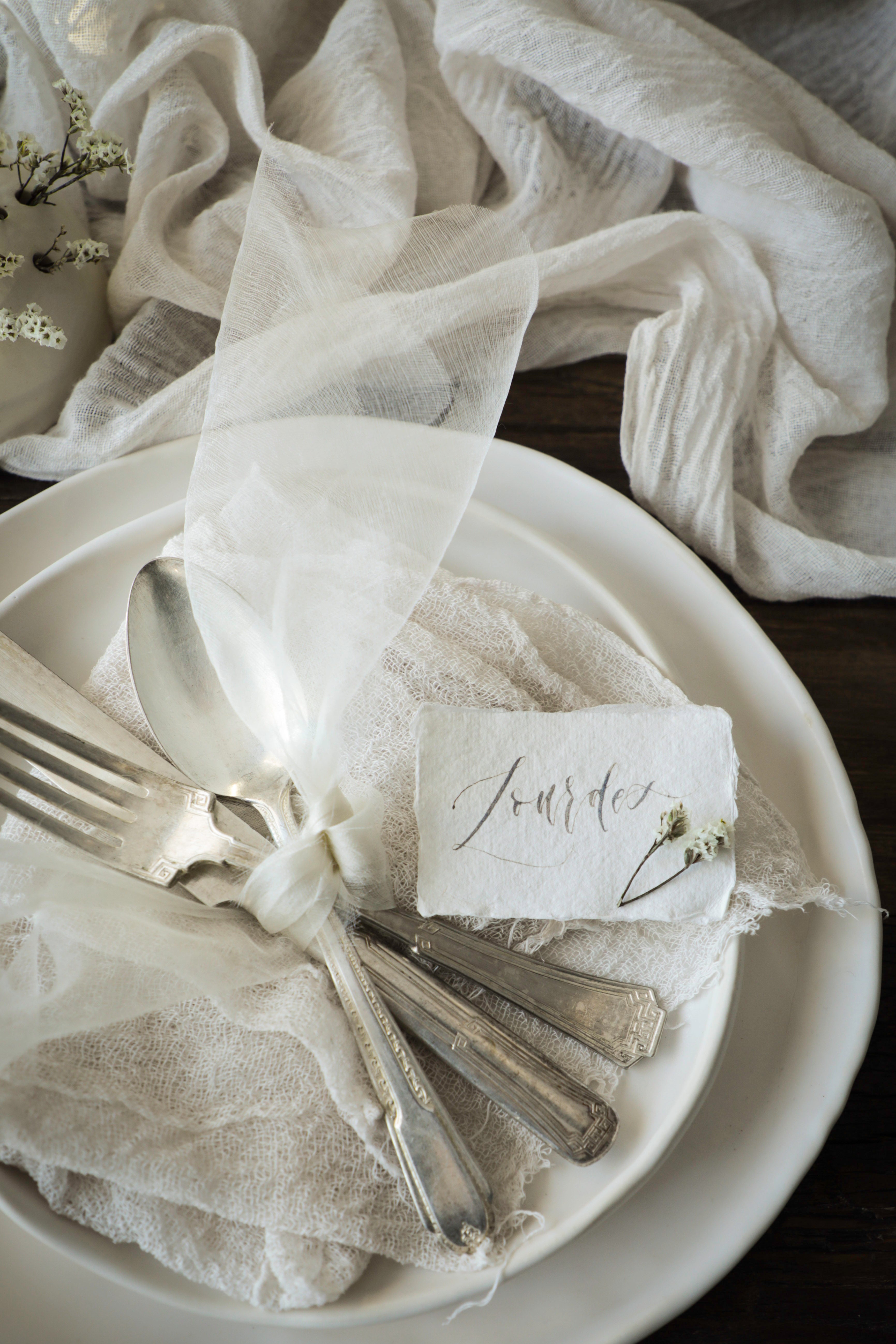 A styled place setting on a white plate with silver-toned flatware, a small white napkin, and a paper place card reading 'Journée,' wrapped in sheer white fabric against a dark wood background with draped white cloth.