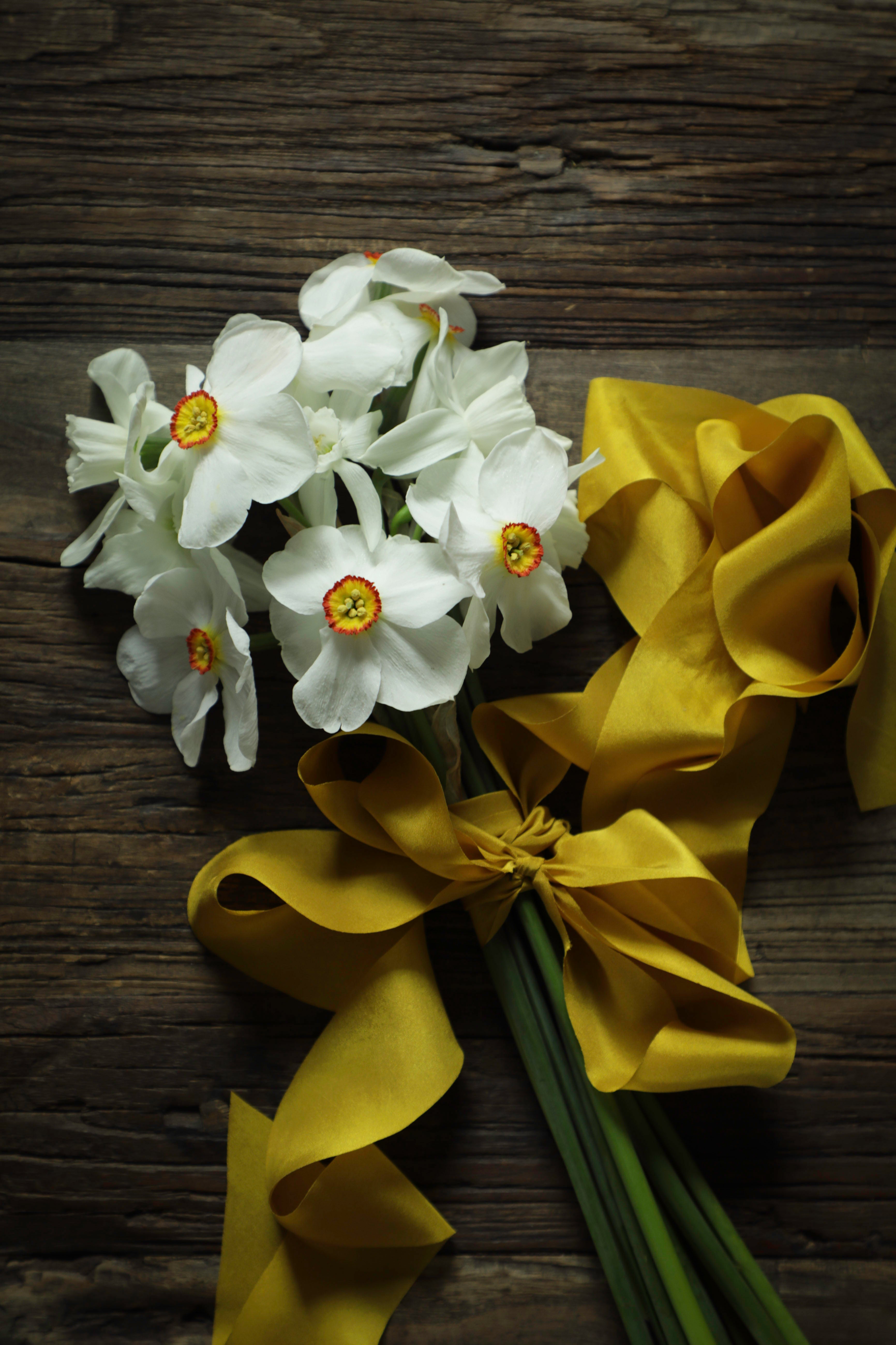 A bouquet of white daffodils with yellow-orange centers tied with a wide yellow satin ribbon, arranged on a dark rustic wooden surface.