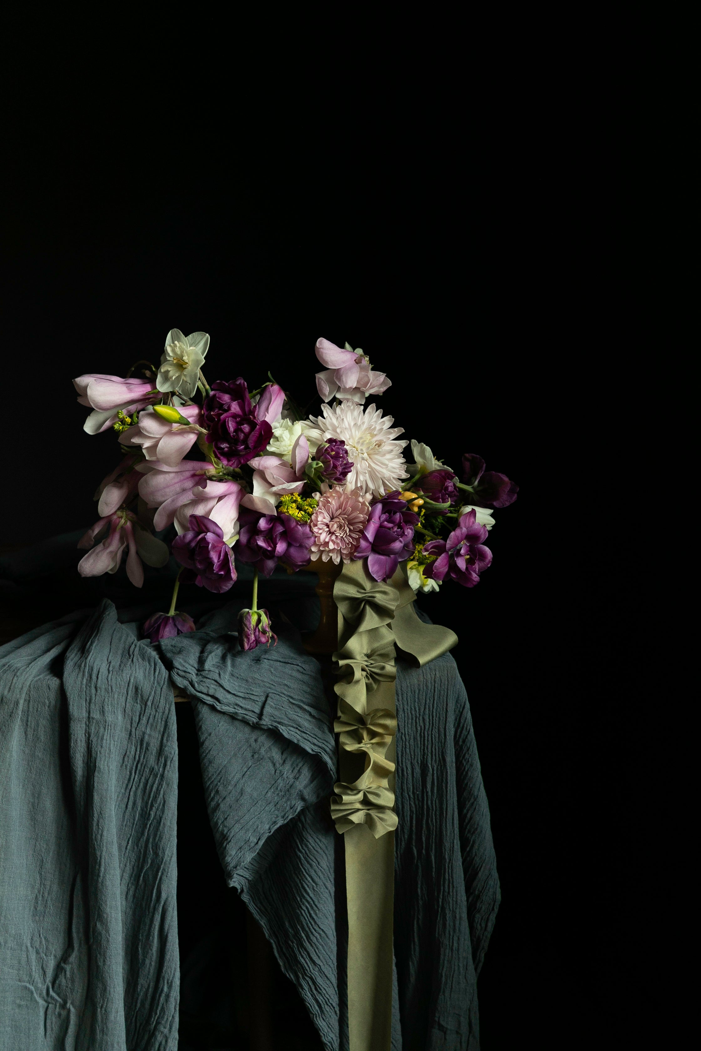 A floral arrangement with purple, pink, and white flowers draped over dark teal and olive-green textured fabric against a solid black background.