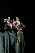 A floral arrangement with purple, pink, and white flowers draped over dark teal and olive-green textured fabric against a solid black background.