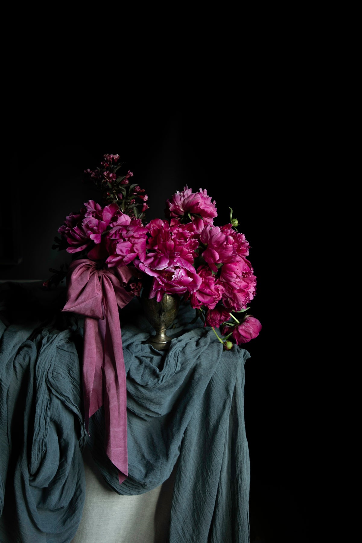 A bouquet of vibrant magenta peonies in a small metallic vase, draped with a dusty blue-gray silk scarf against a solid black background.