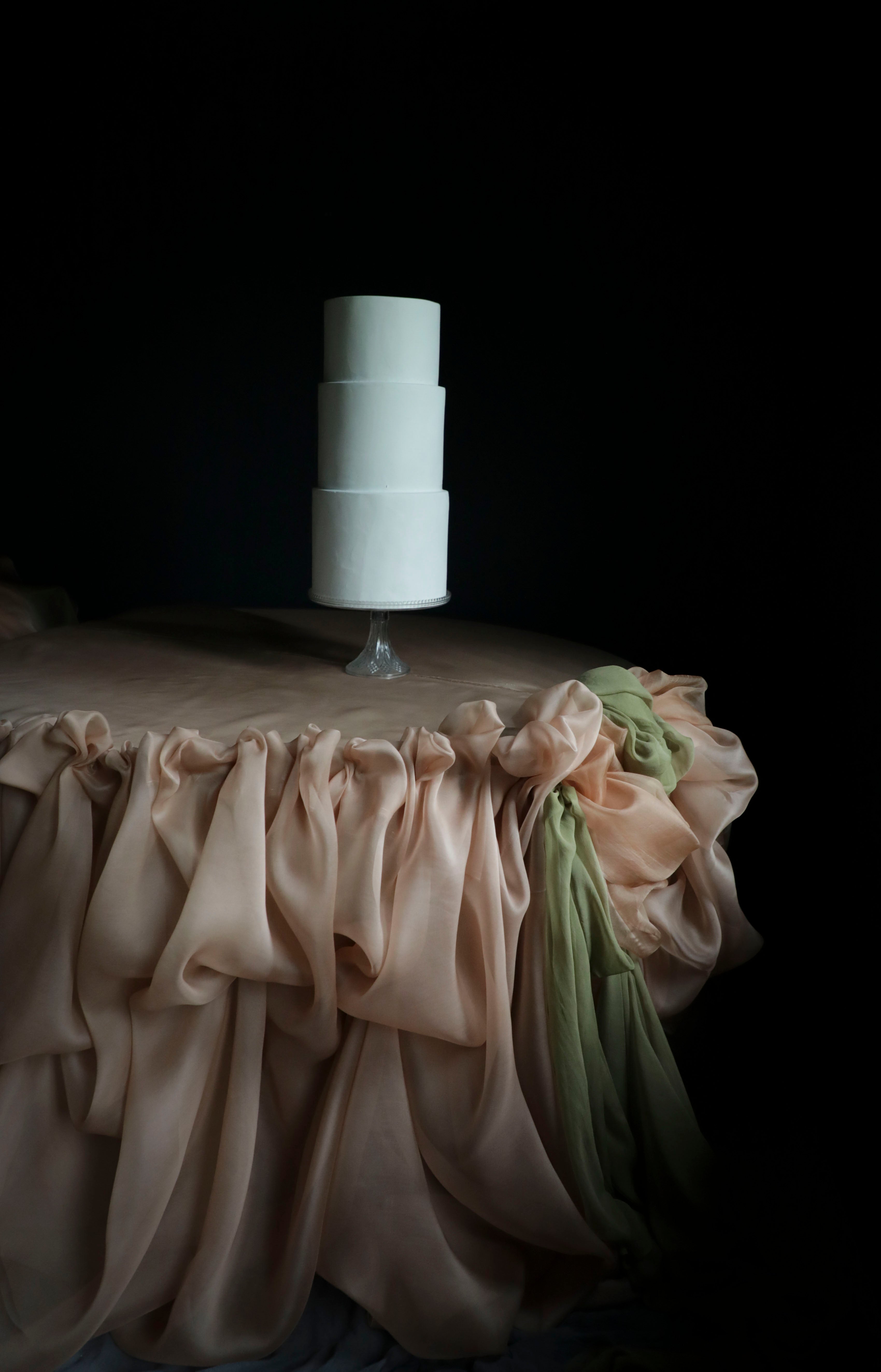 A three-tier white cake on a clear stand, set against a dark background with a draped champagne tablecloth and a green fabric accent.