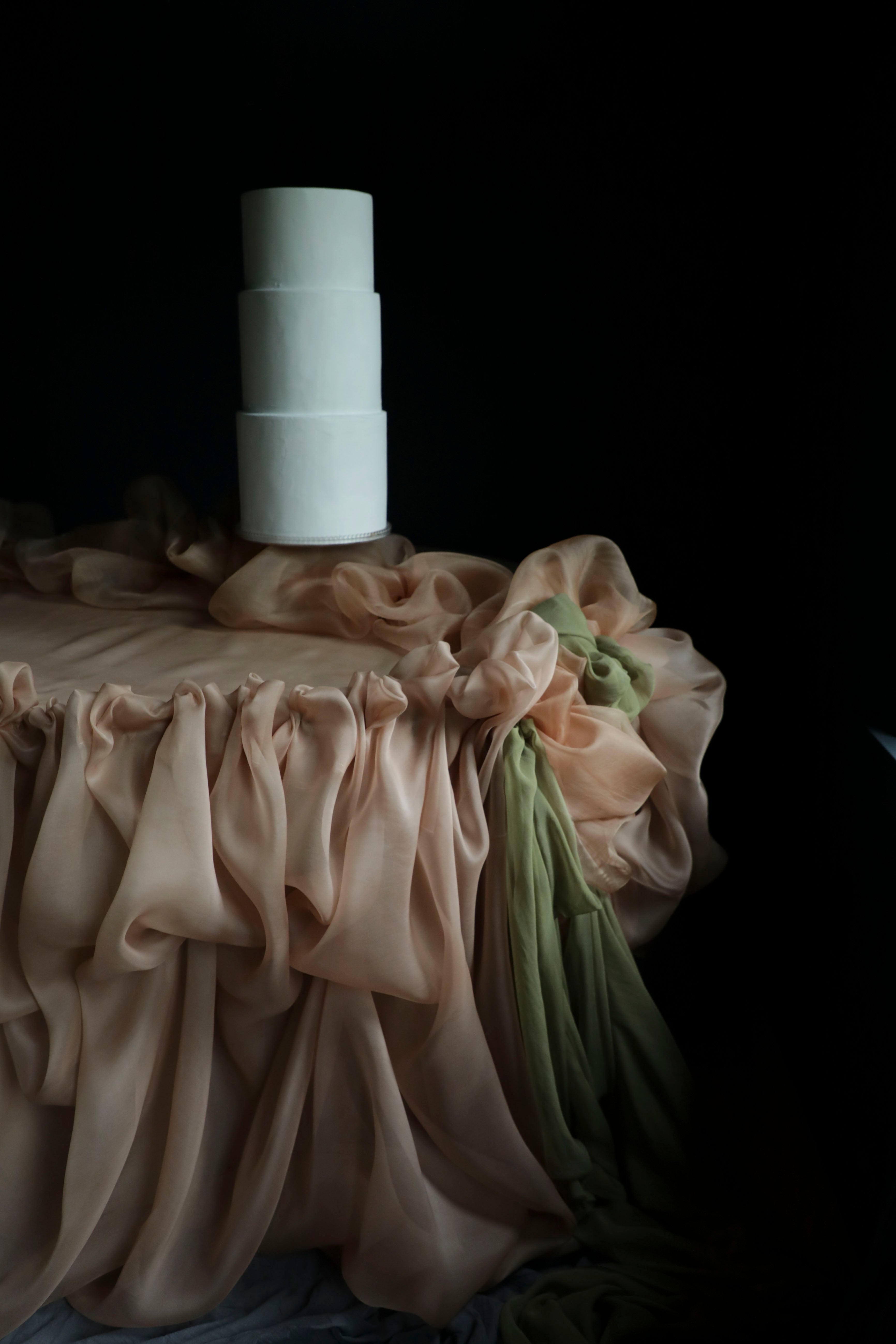 A white wedding cake on a draped surface covered in soft blush silk fabric with a green section, set against a very dark background.