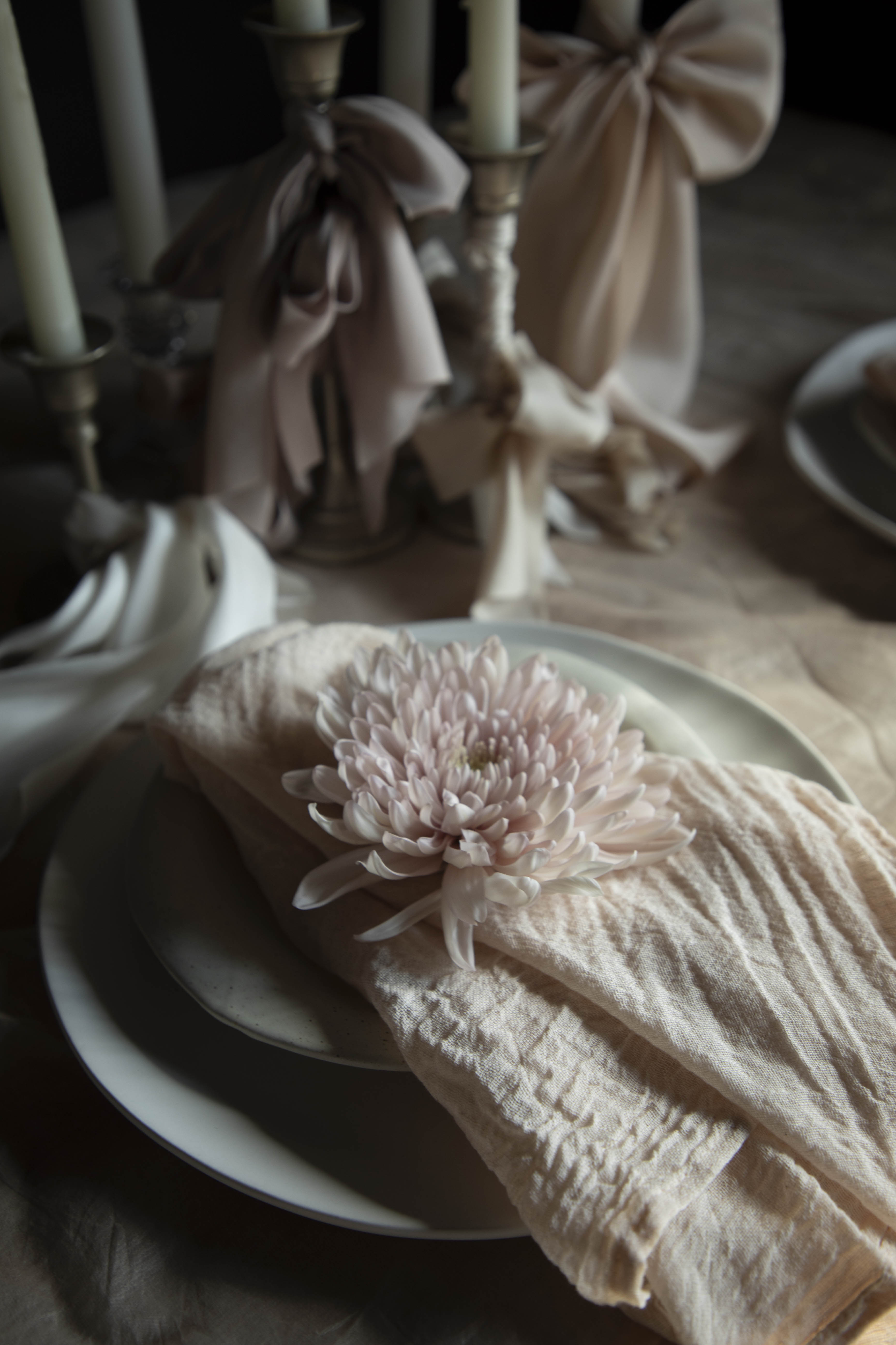 Neatly folded beige napkin with a decorative flower on a white plate, set against a blurred background of candles and bows.