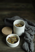 Two small white ceramic cups filled with loose herbal tea, placed on a dark wooden surface with a gray cloth and a cork lid nearby.