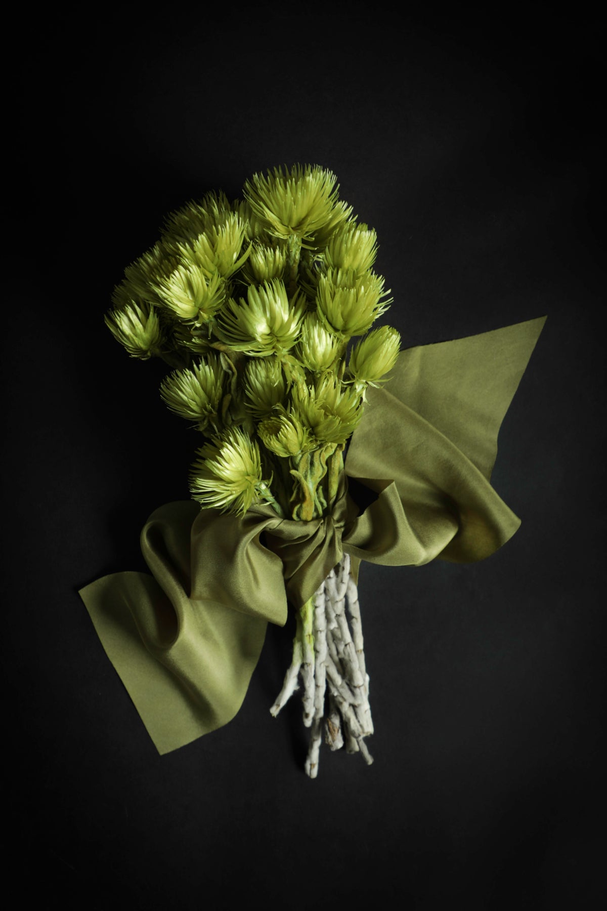 A bouquet of light green dried flowers wrapped in olive green paper and tied with a matching ribbon, set against a solid black background.
