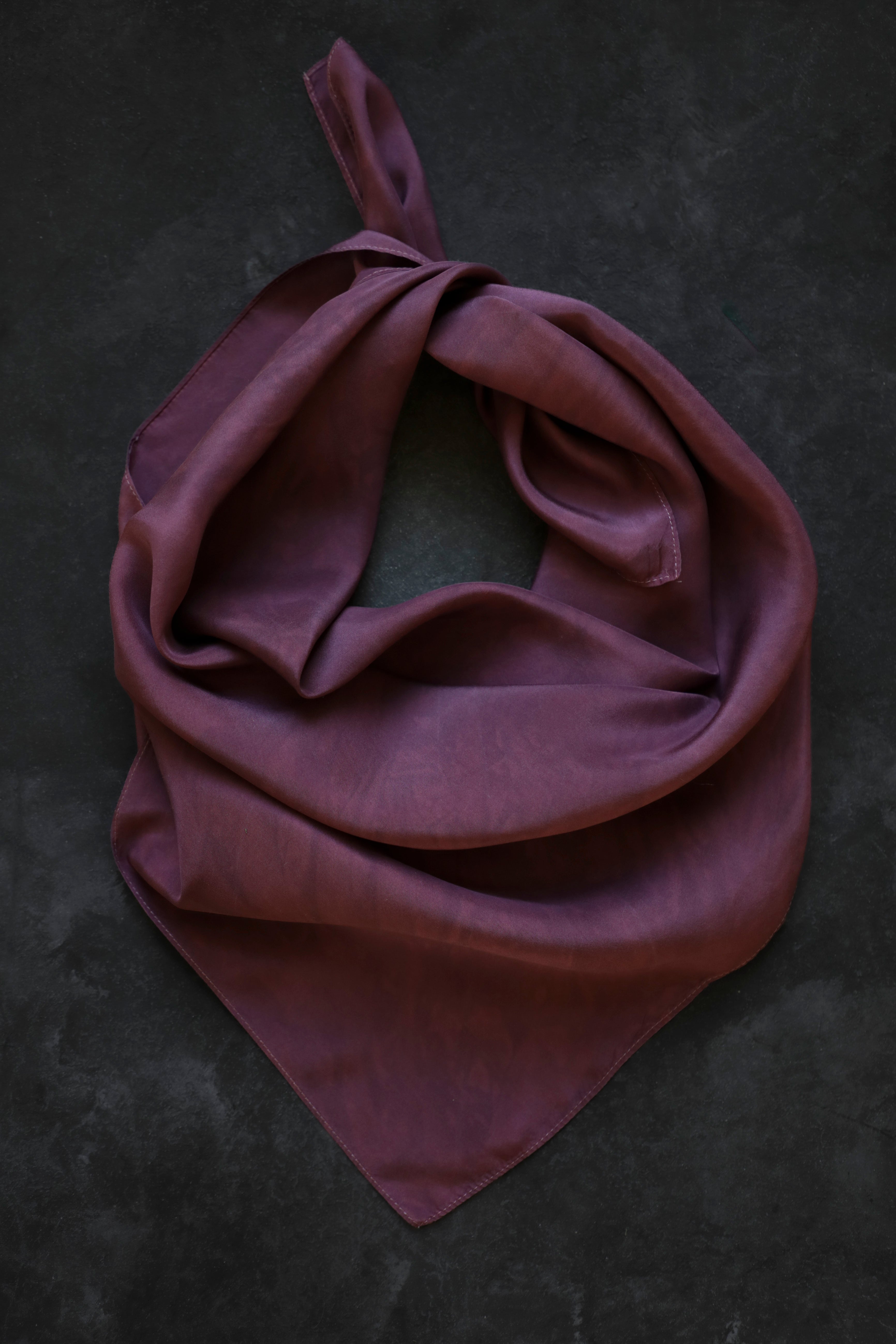 A mauve-purple silk scarf tied in a loose knot, displayed on a dark textured background.