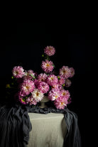 A bouquet of pink and white dahlia flowers arranged in a ceramic vase on a draped with dark gray textiles on a table against a solid black background.