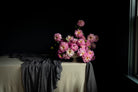A bouquet of pink and white dahlia flowers arranged in a ceramic vase on a draped table, set against a dark black background with a light-colored linen fabric covering the table.