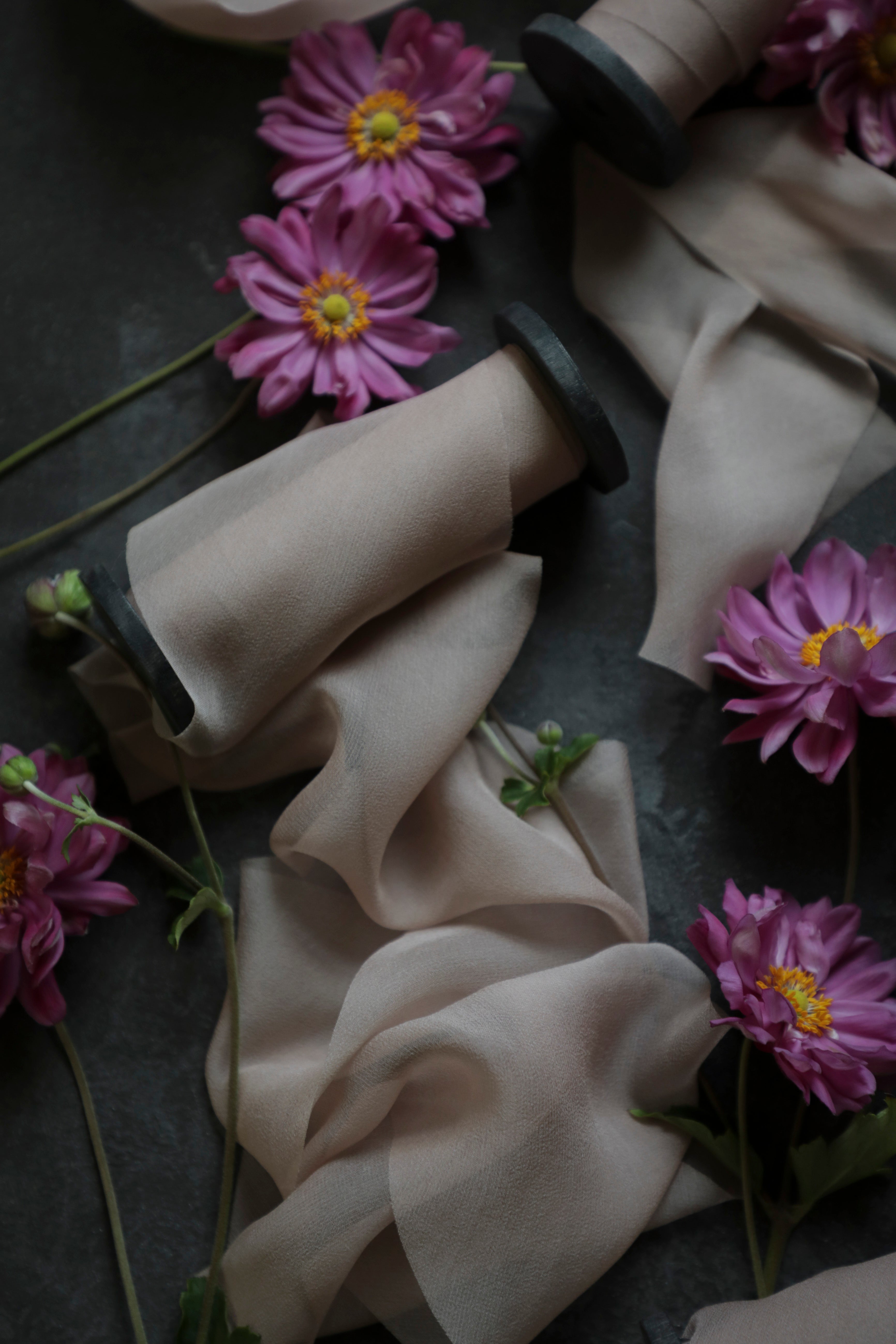 A styled flat lay of sheer mauve ribbons on black wooden spools is arranged on a dark surface, with scattered mauve flowers with yellow centers.